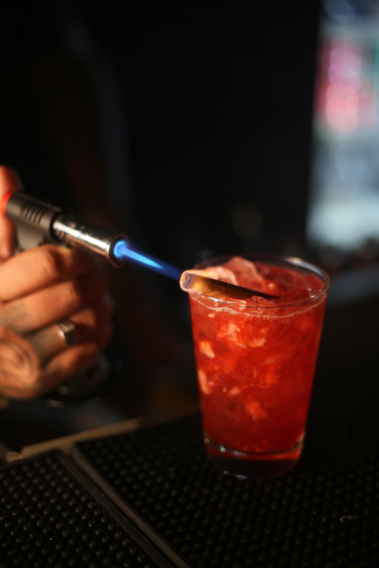 Person using a blowtorch to caramelize the top of a red cocktail drink in a tall glass.