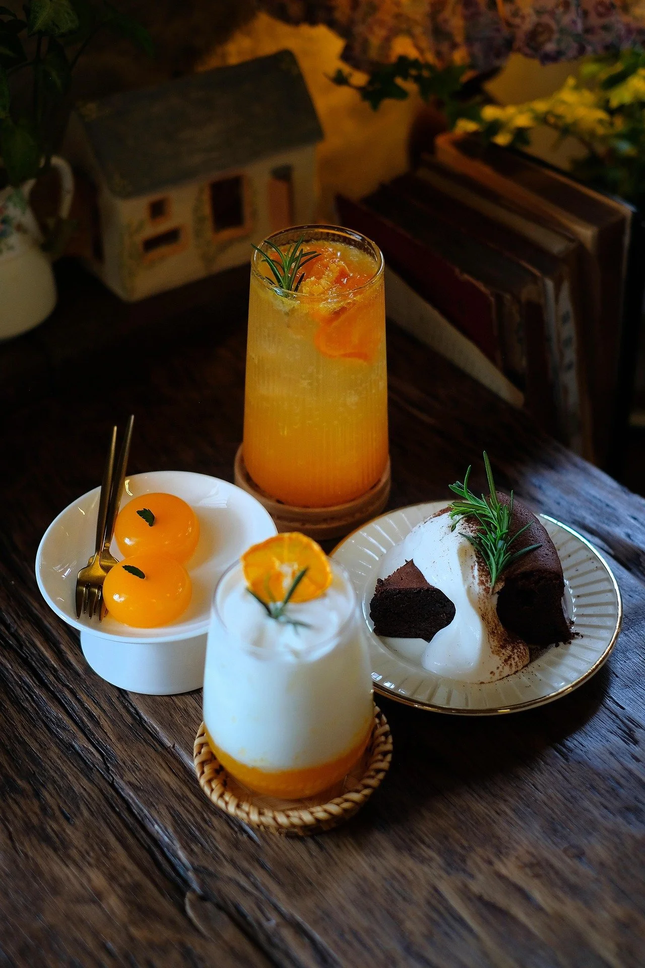 A table with various desserts and drinks including a citrus cocktail, an orange-dessert topped with cream and a sprig of rosemary, a chocolate cake with cream, and a plate with two yellow gel-like balls topped with rosemary, set on a rustic wooden table.