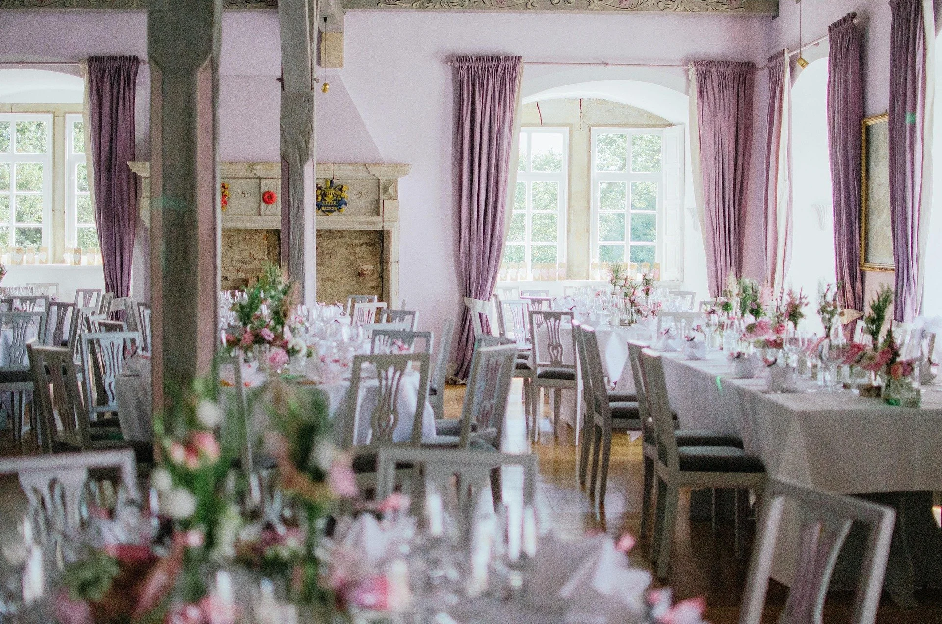 Elegant banquet hall decorated for a wedding with long tables covered in white tablecloths, pink floral centerpieces, and chairs with patterned backs. Large windows with pink curtains allow natural light into the room, which features a stone fireplac