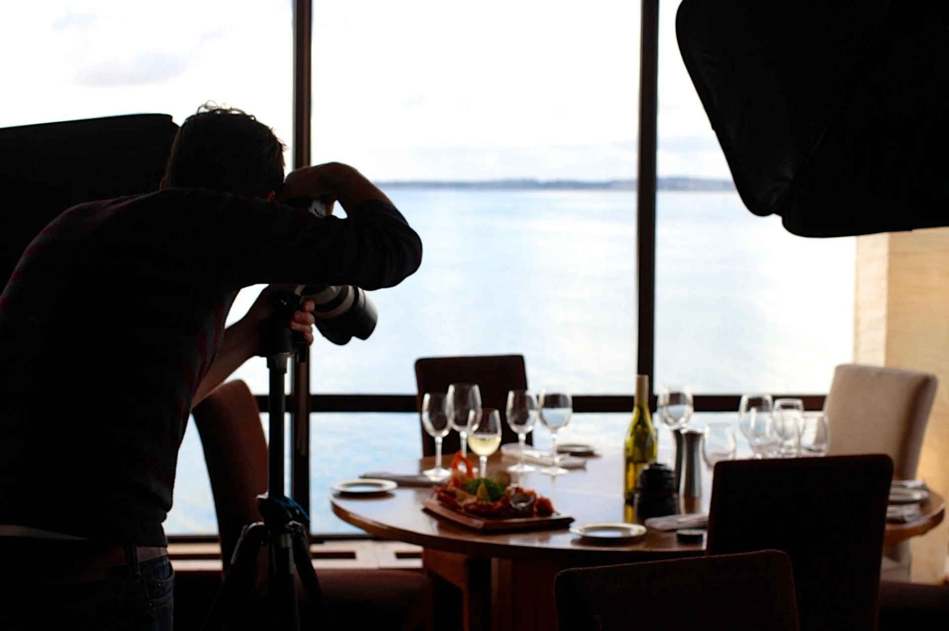 Photographer with camera taking pictures of a dining setup near water, with wine bottle and glasses on the table, in a room with large windows.
