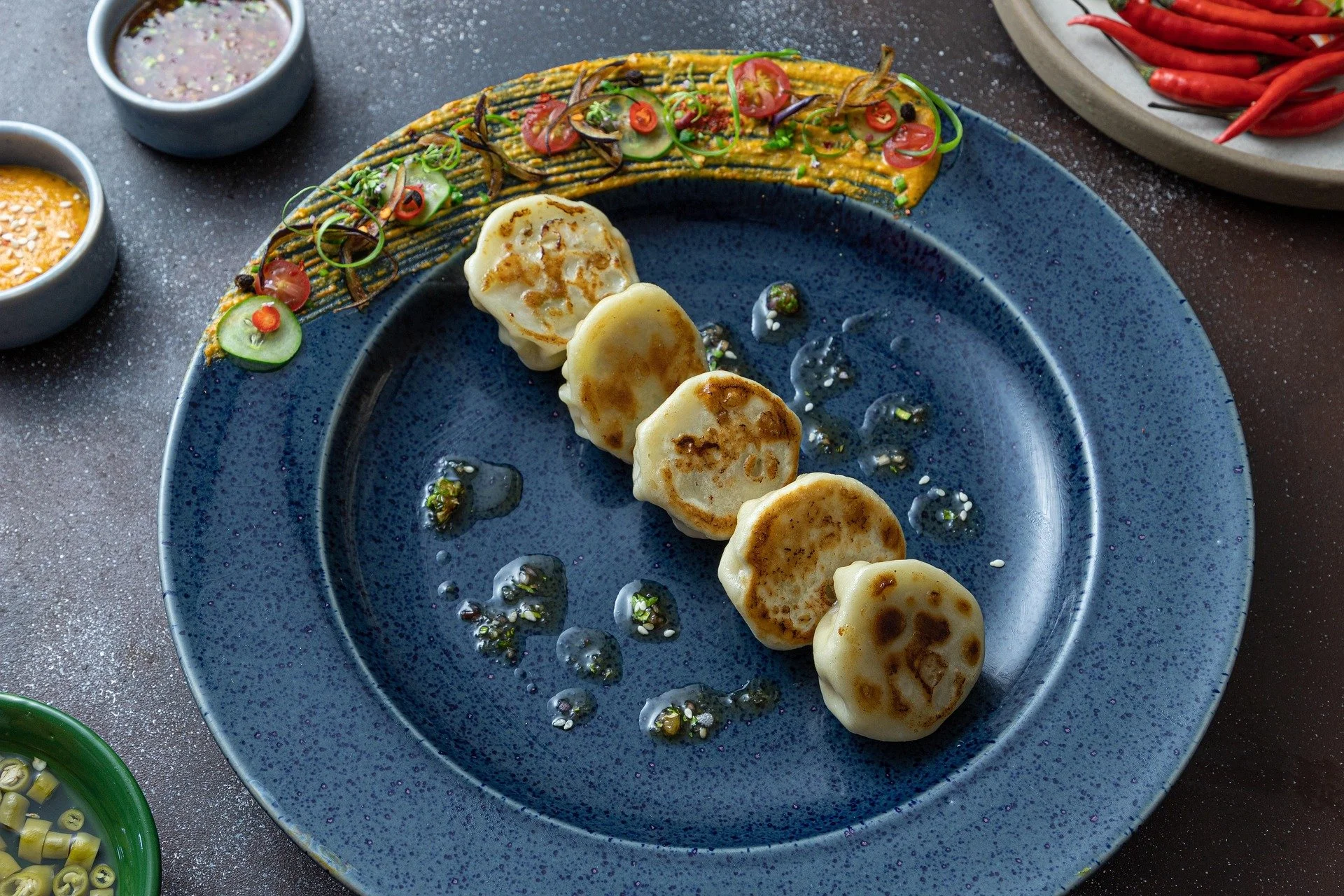 Five pan-fried dumplings on a blue speckled plate with soy sauce drops and scallions, garnished with a colorful vegetable drizzle and small cups of dipping sauces, surrounded by bowls of chili peppers, sauce, and chopped green onions.