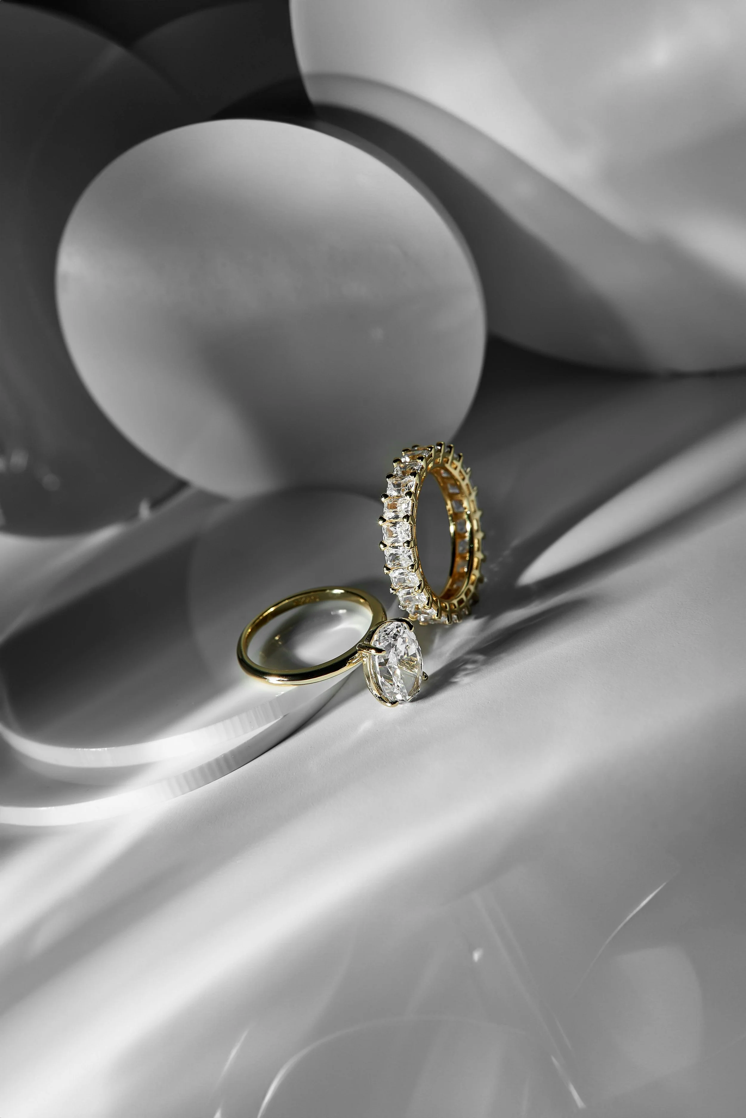 Three jewelry rings, including a gold band, a diamond engagement ring, and a gold ring with embedded diamonds, are placed on a reflective satin surface with a modern abstract sculpture background.