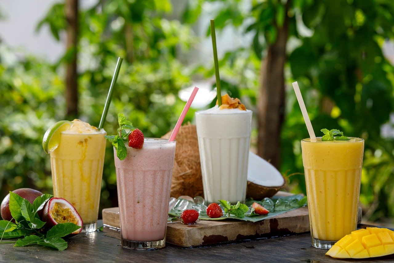 Four tropical smoothies in glasses with straws, garnished with fruits and mint, set outdoors with green trees in the background.