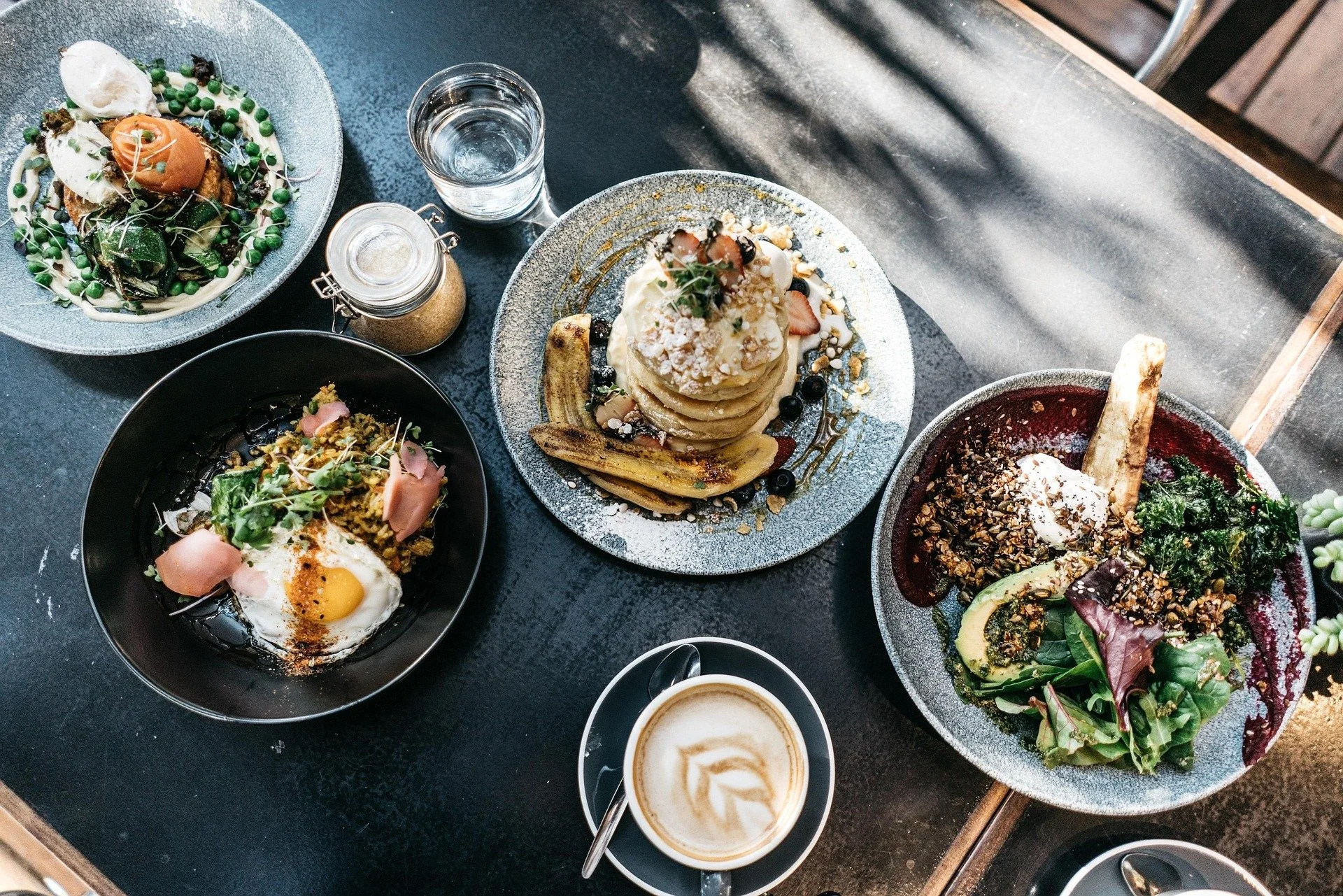 A top-down view of a table with four plates of food, a cup of coffee, a glass of water, and condiments. The dishes include salad, pancakes topped with fruit and syrup, a rice bowl with an egg, and a dessert with a cookie and whipped cream.