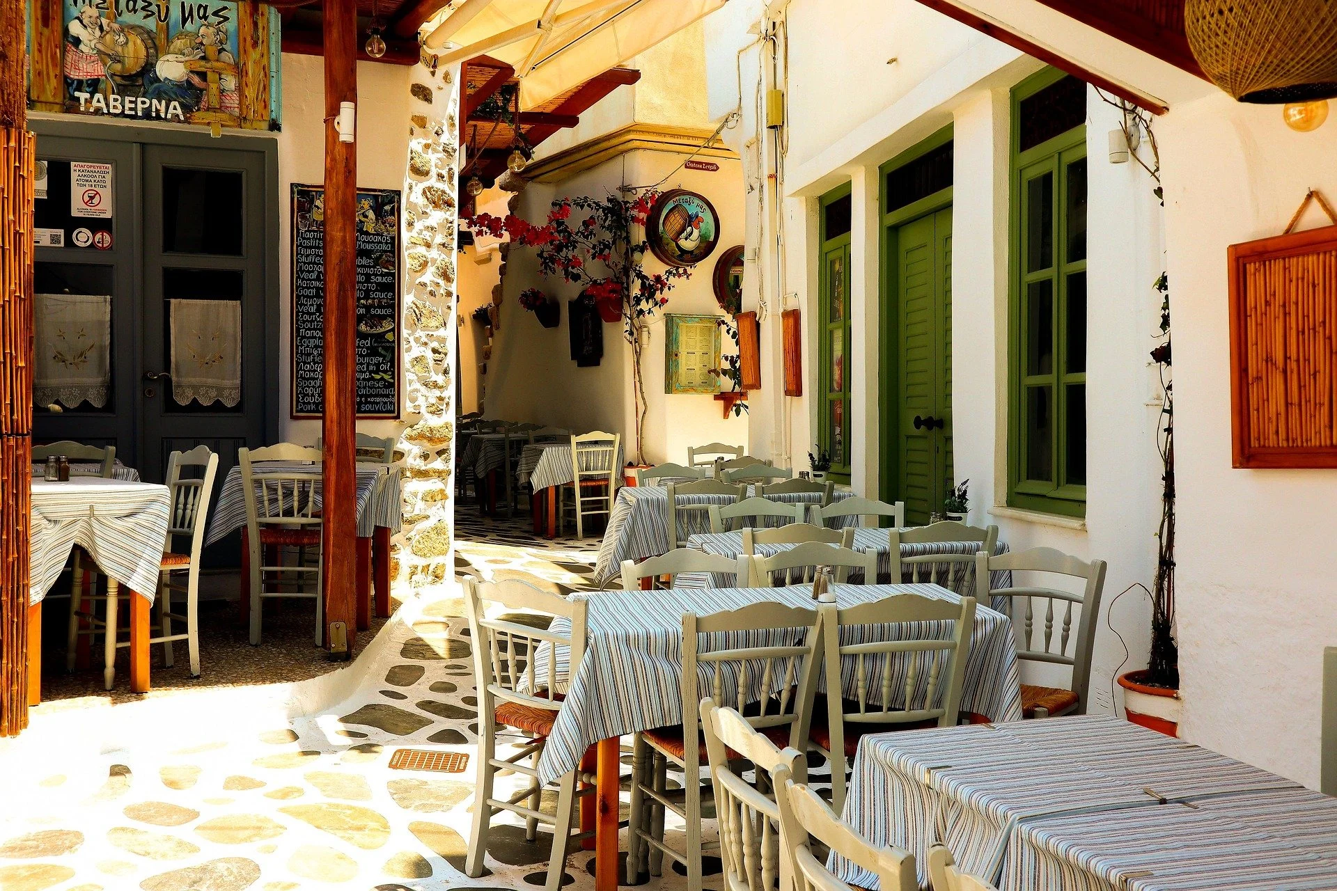 Outdoor seating area of a Greek taverna with striped tablecloths, white wooden chairs, green-framed windows, and decorative wall hangings.