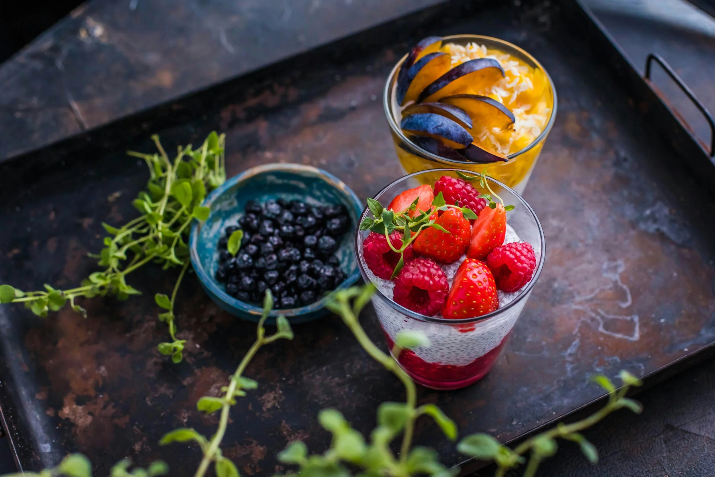 Two glasses of fruit desserts, one with strawberries and raspberries, the other with sliced peaches and purple plums, on a dark metal tray with a small bowl of blueberries and some green foliage.