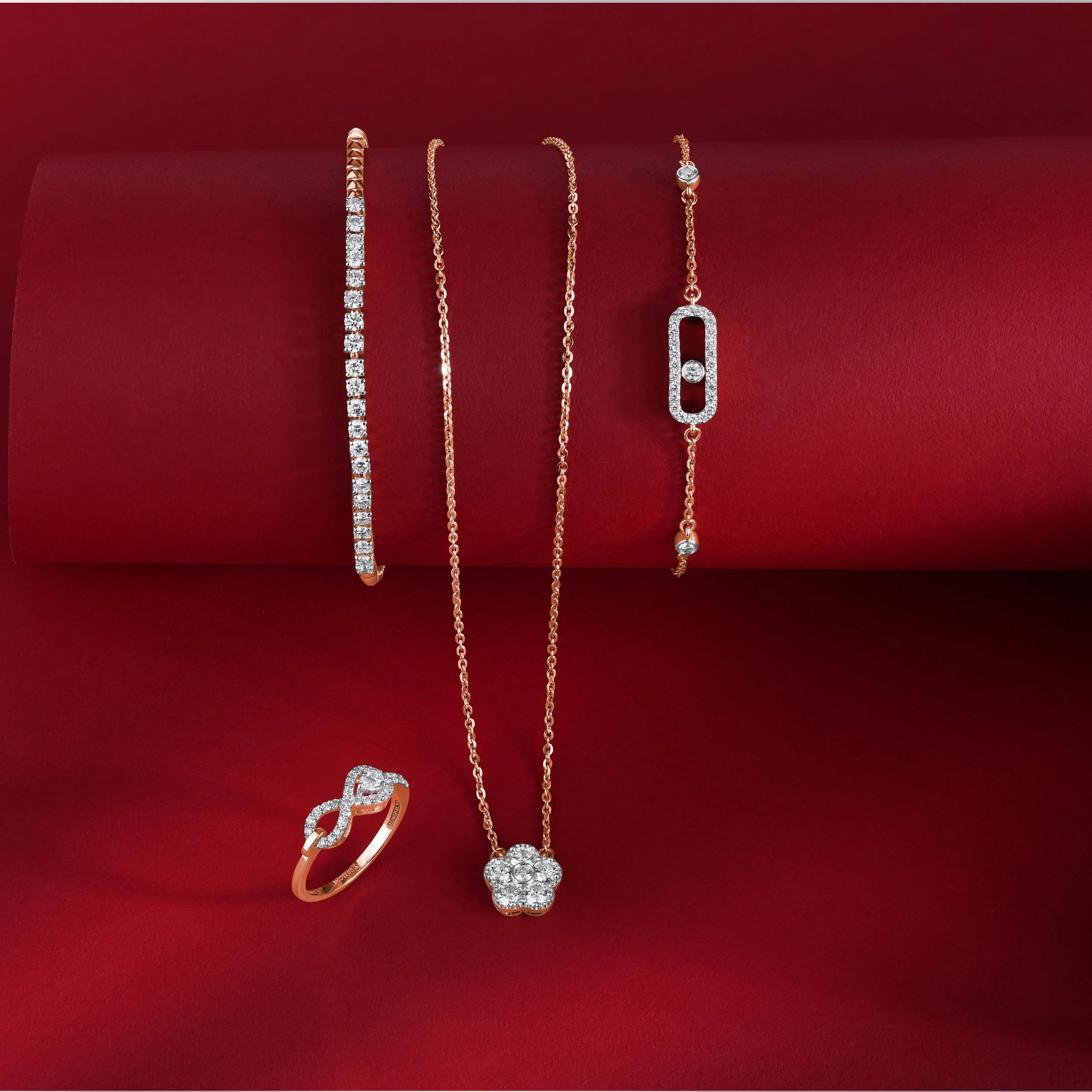 Set of jewelry including a diamond bracelet, a gold chain necklace with a flower-shaped pendant, a gold chain bracelet with a rectangular gemstone, and a gold ring with a bow-shaped design, displayed on a red background.