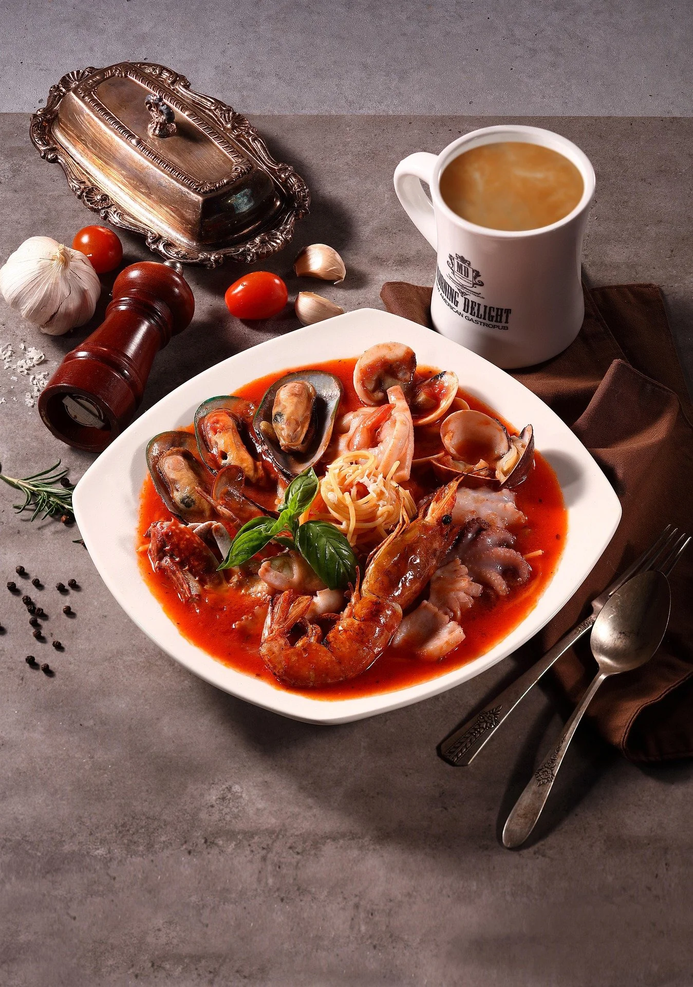 Seafood pasta with shrimp, mussels, and tomato sauce, garnished with basil leaves, served with a cup of soup and surrounded by garlic, cherry tomatoes, and seasoning on a gray surface.