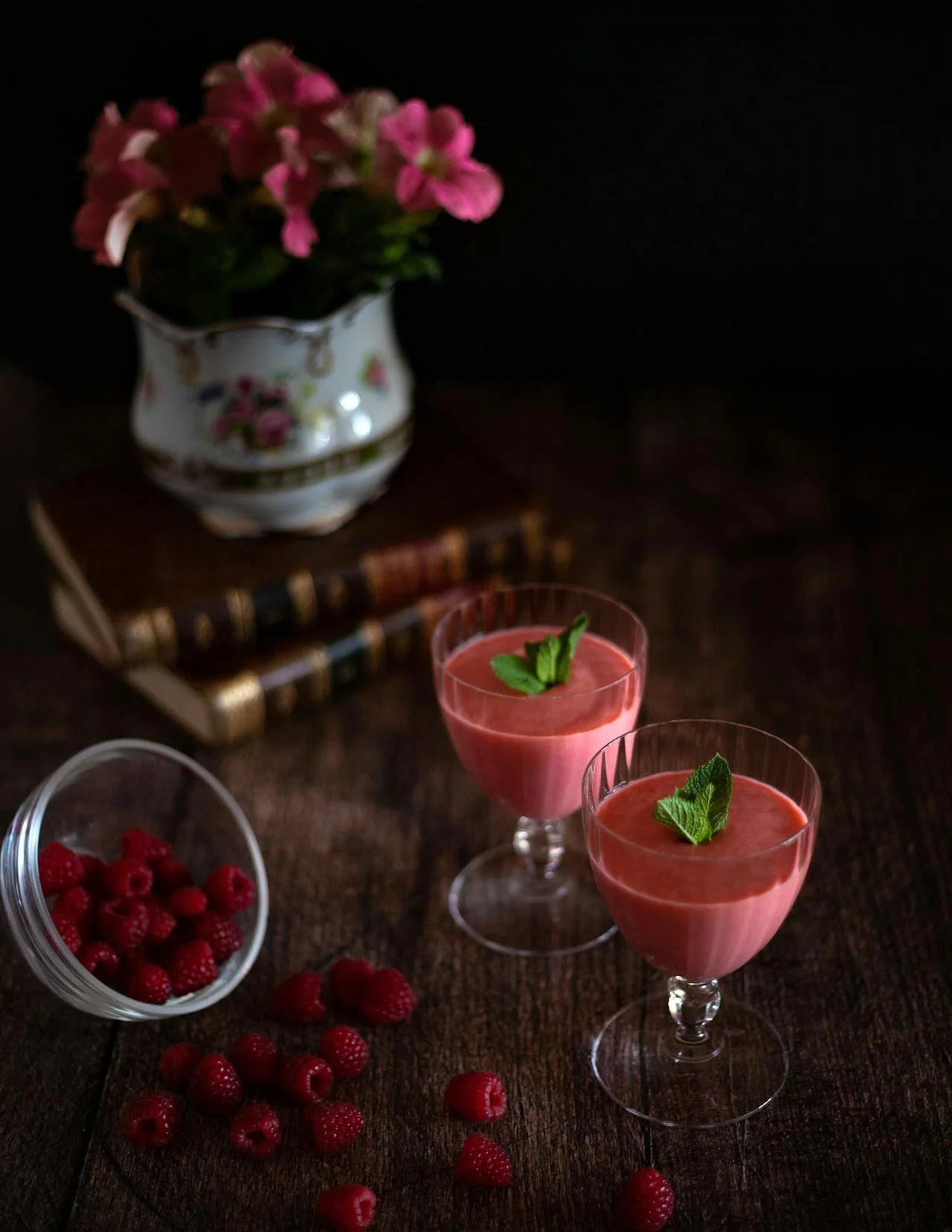 Two glasses of pink raspberry smoothies garnished with green mint leaves on a dark wooden table, with a bowl of fresh raspberries and radishes nearby, and a vase of pink flowers in the background.