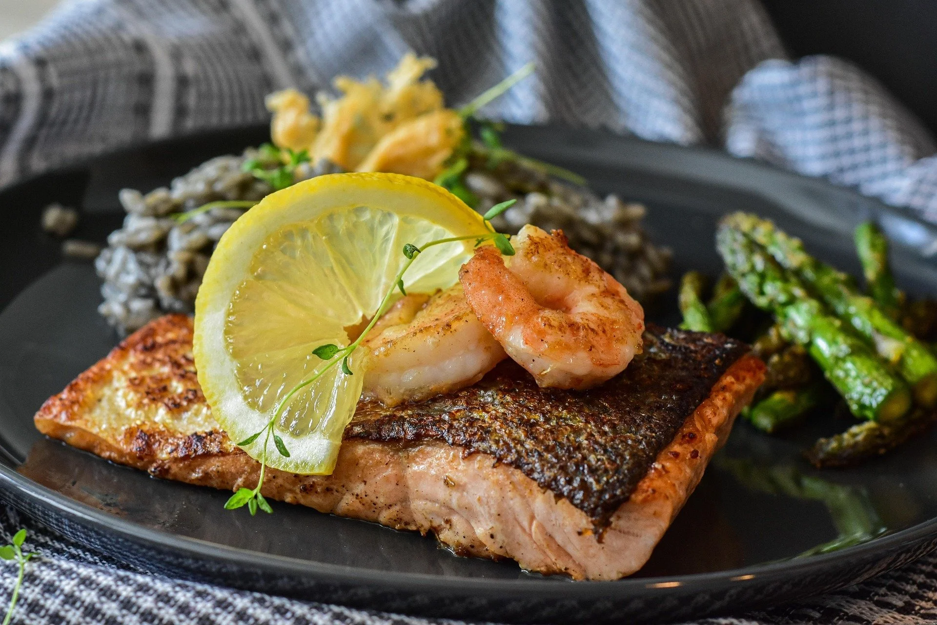 Grilled salmon with shrimp, lemon slice, and microgreens on a black plate, accompanied by sautéed green beans and a side grain dish.
