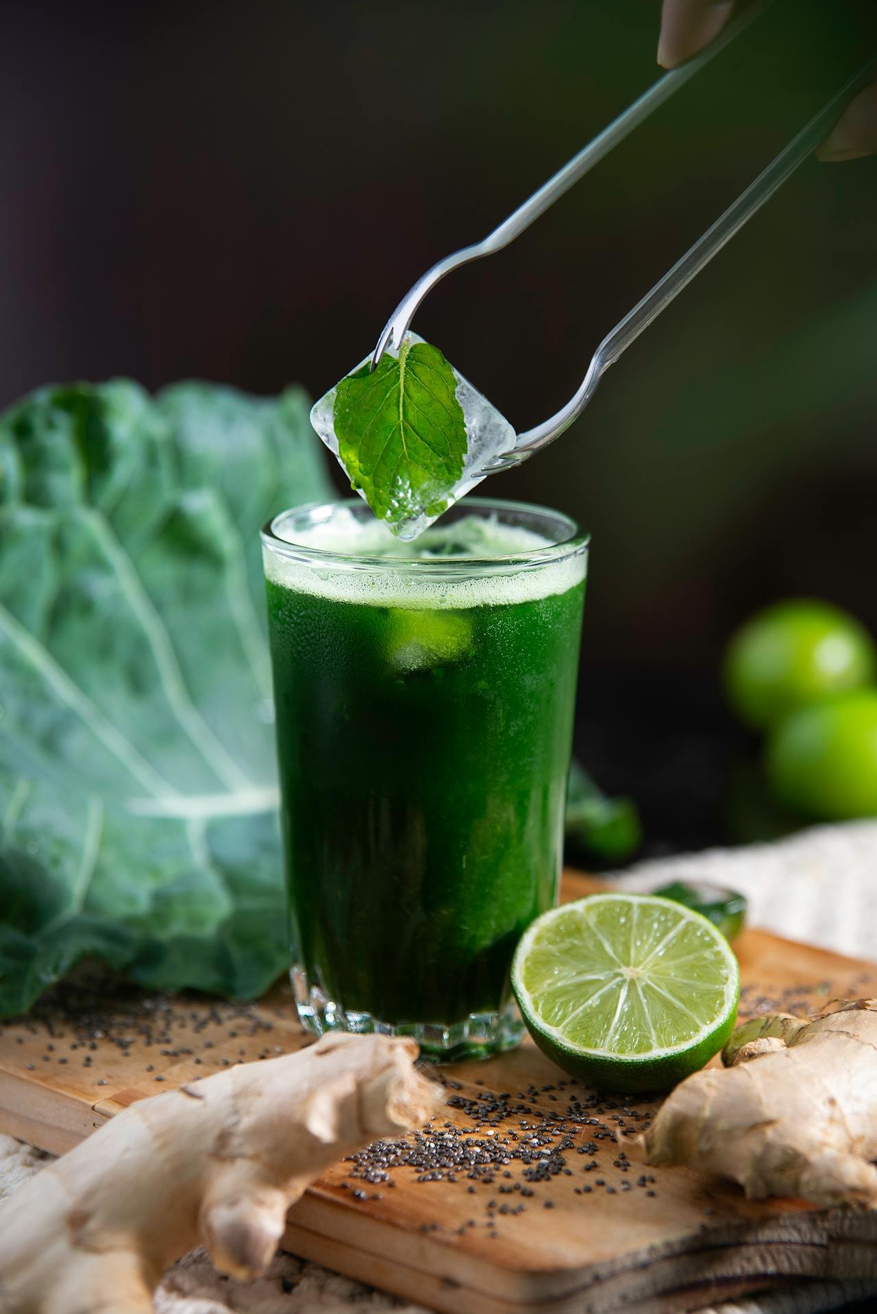 A tall glass of green juice with lime, ginger, and leafy greens, with a metal tong holding an ice cube and a leaf above the glass, surrounded by ingredients like ginger, lime, and black seeds on a wooden cutting board.