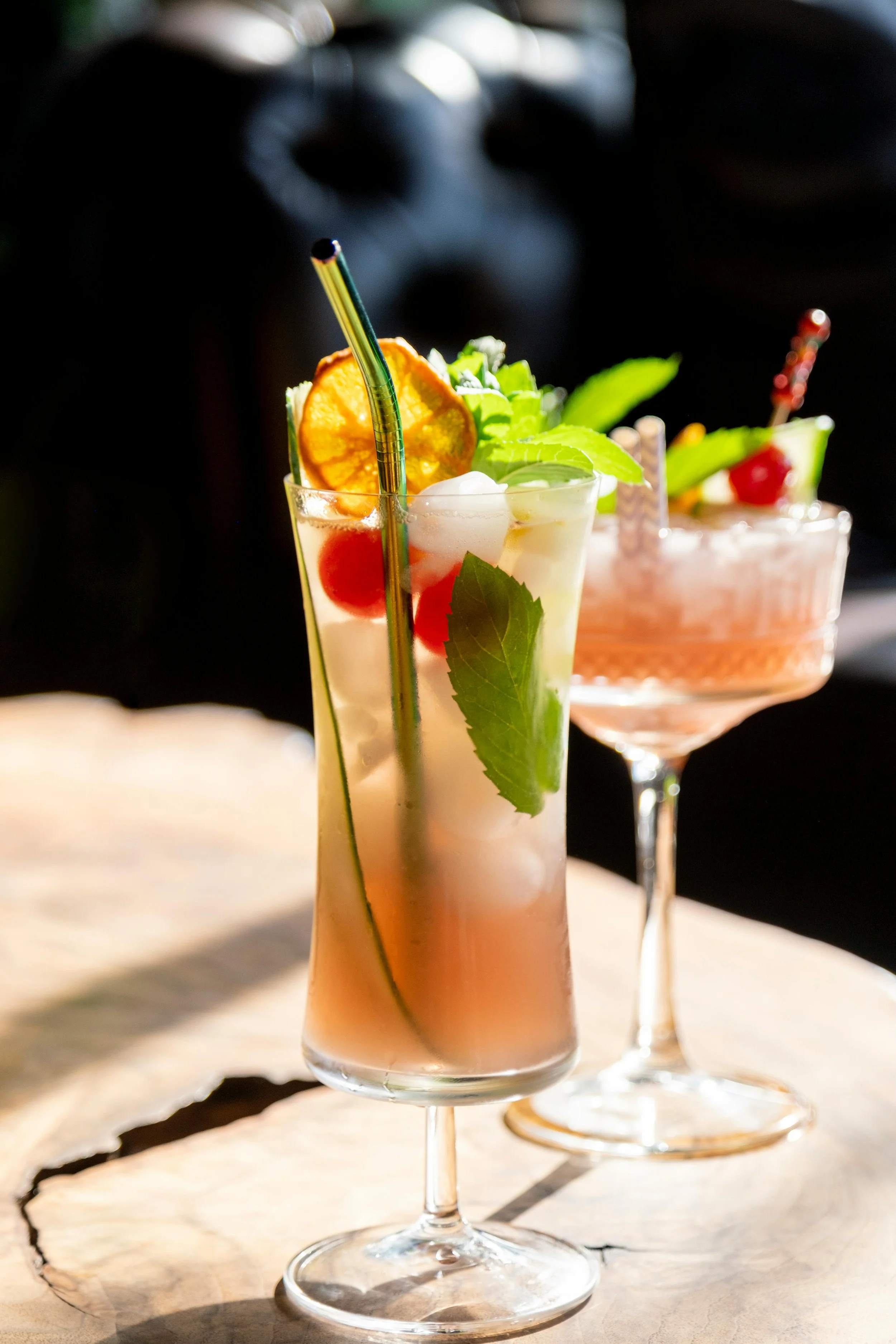 Two colorful cocktails with garnishes, one in a tall glass with a straw and citrus slice, and the other in a stemmed glass with a cherry and mint, on a wooden surface.