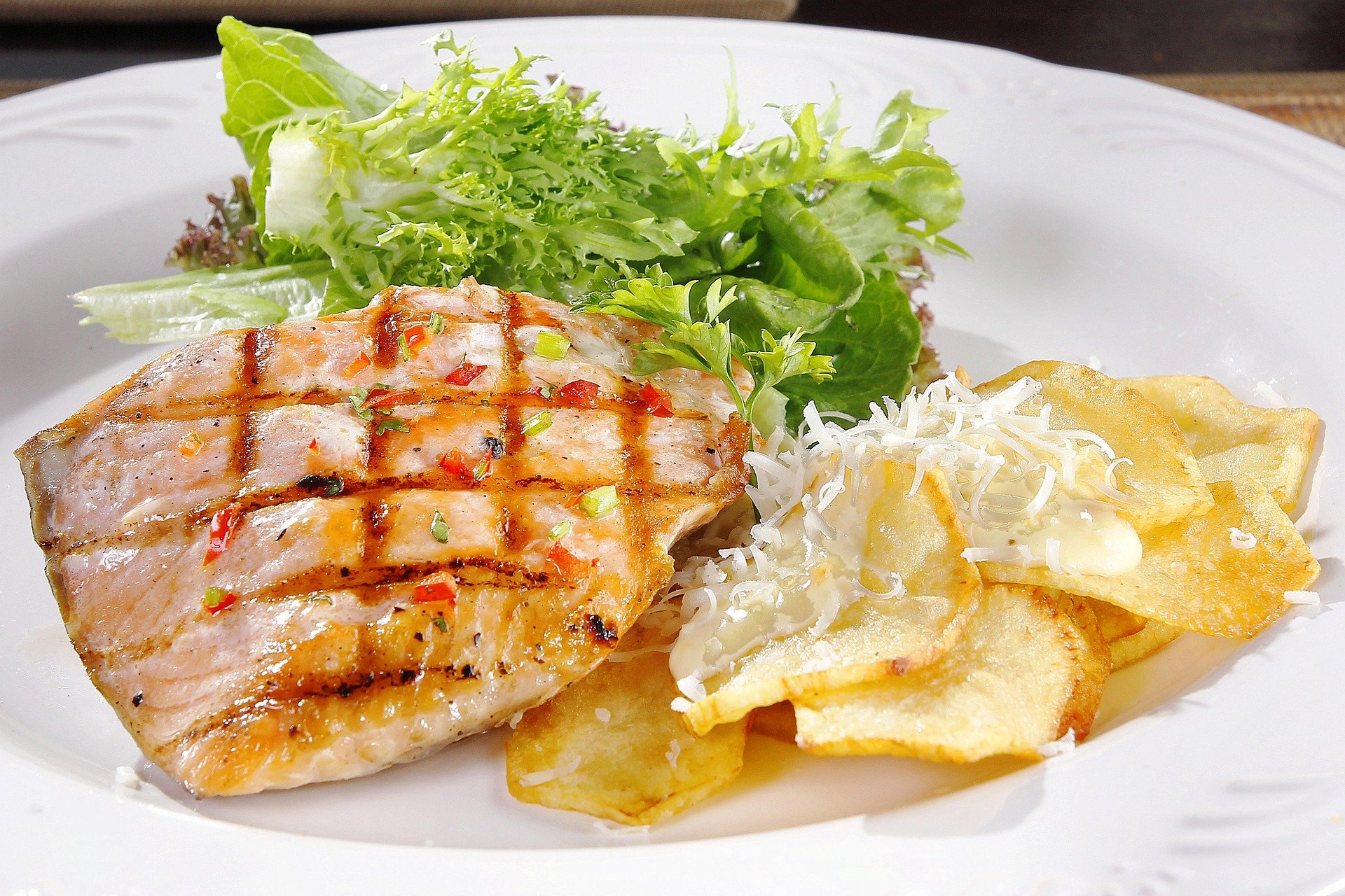 Grilled chicken breast with salad greens, potato chips, grated cheese, and creamy sauce on a white plate.