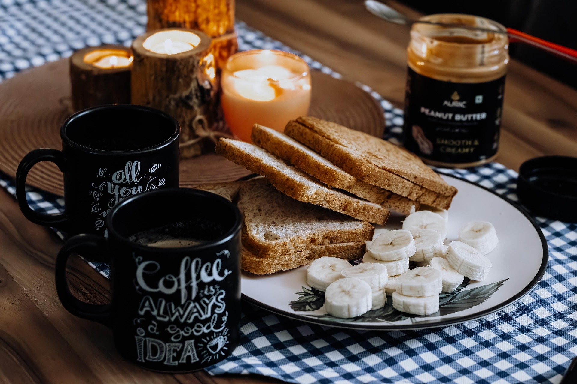 Breakfast scene with sliced bread, banana slices, and a jar of peanut butter on a decorative plate. Two black mugs with coffee, decorated with white writing, sit on a checkered tablecloth. Candles and wooden decorations add warm ambiance.