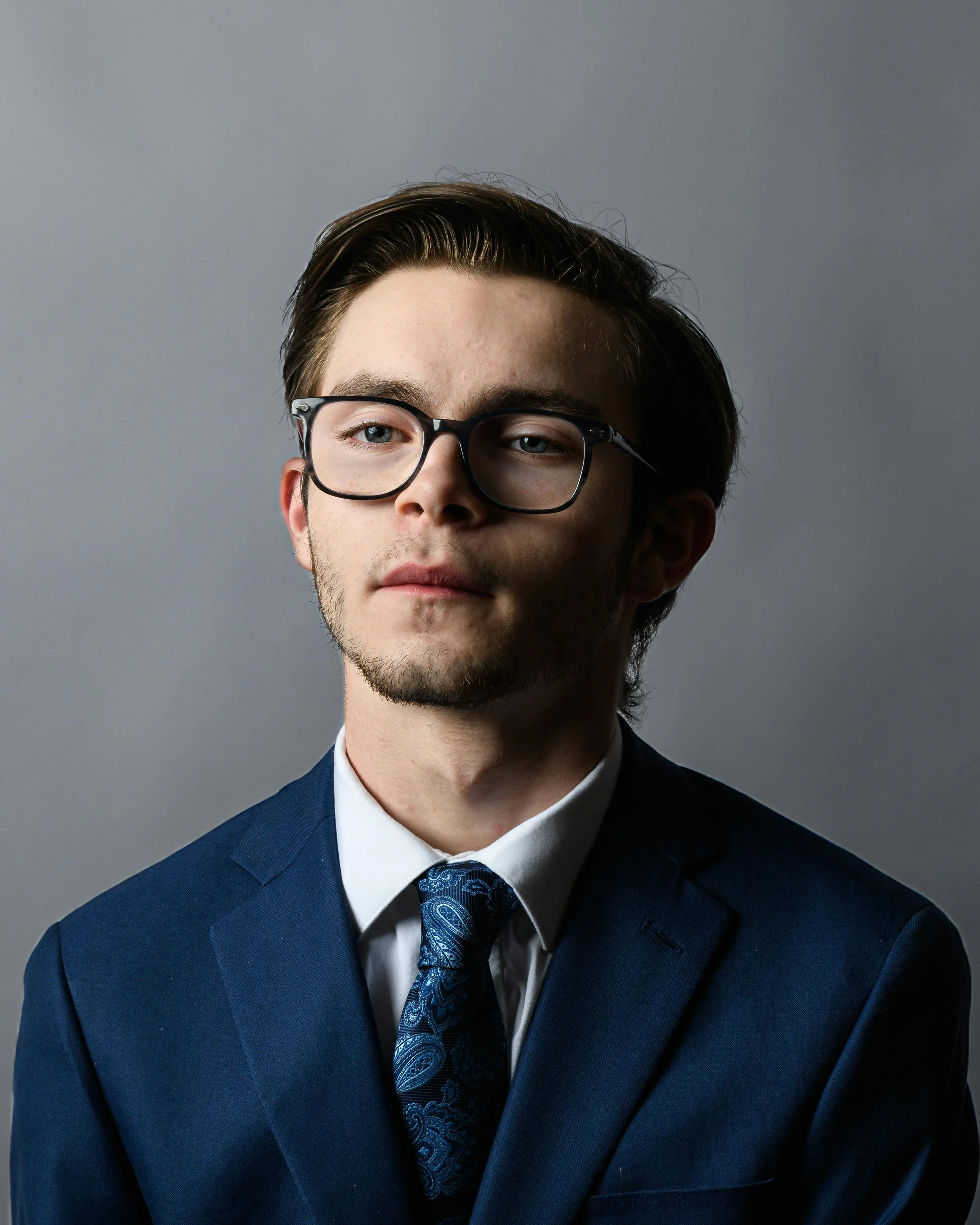 Portrait of a young man with dark hair, glasses, a beard, wearing a blue suit, white shirt, and blue paisley tie, against a plain gray background.