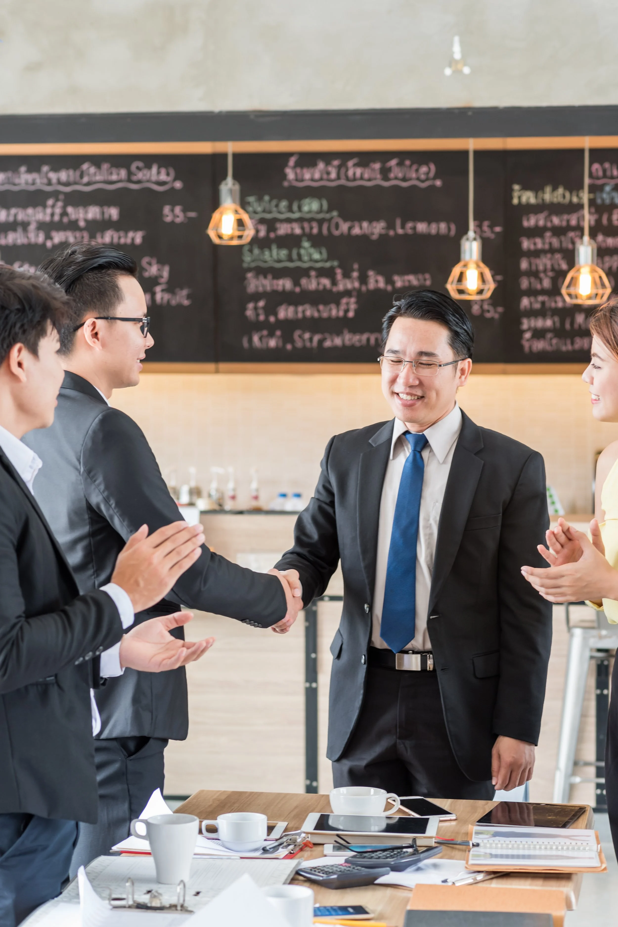 Business people in formal attire exchanging a handshake in a cafe or restaurant, with a chalkboard menu in the background.