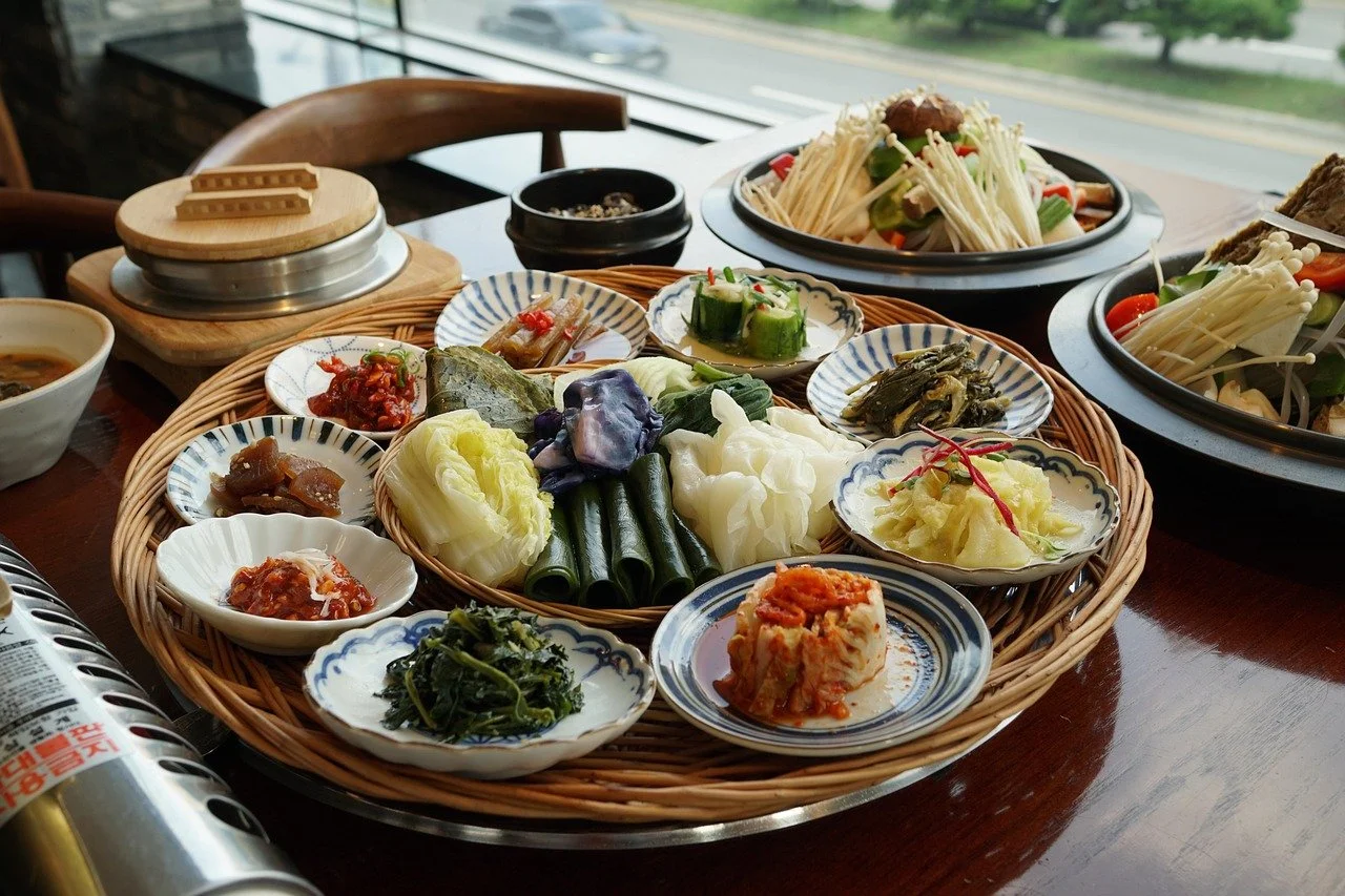 A variety of Korean side dishes arranged on a woven tray, with some dishes in small bowls and others on plates, along with two hot pots of soup and a hot plate of vegetables. The background shows a window and a road outside.