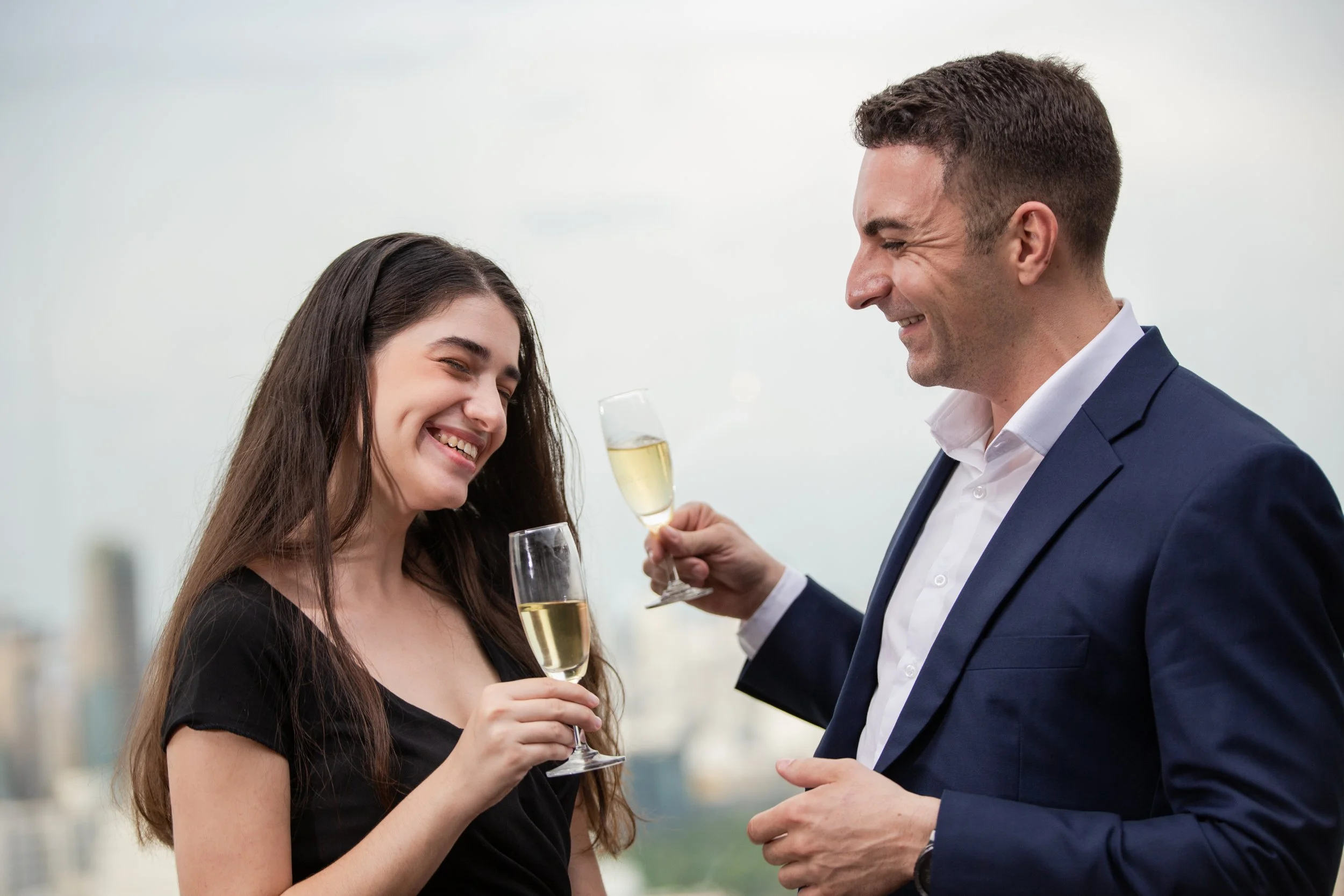 A man and woman celebrating with champagne glasses, both smiling and dressed in formal attire, against a city skyline background.