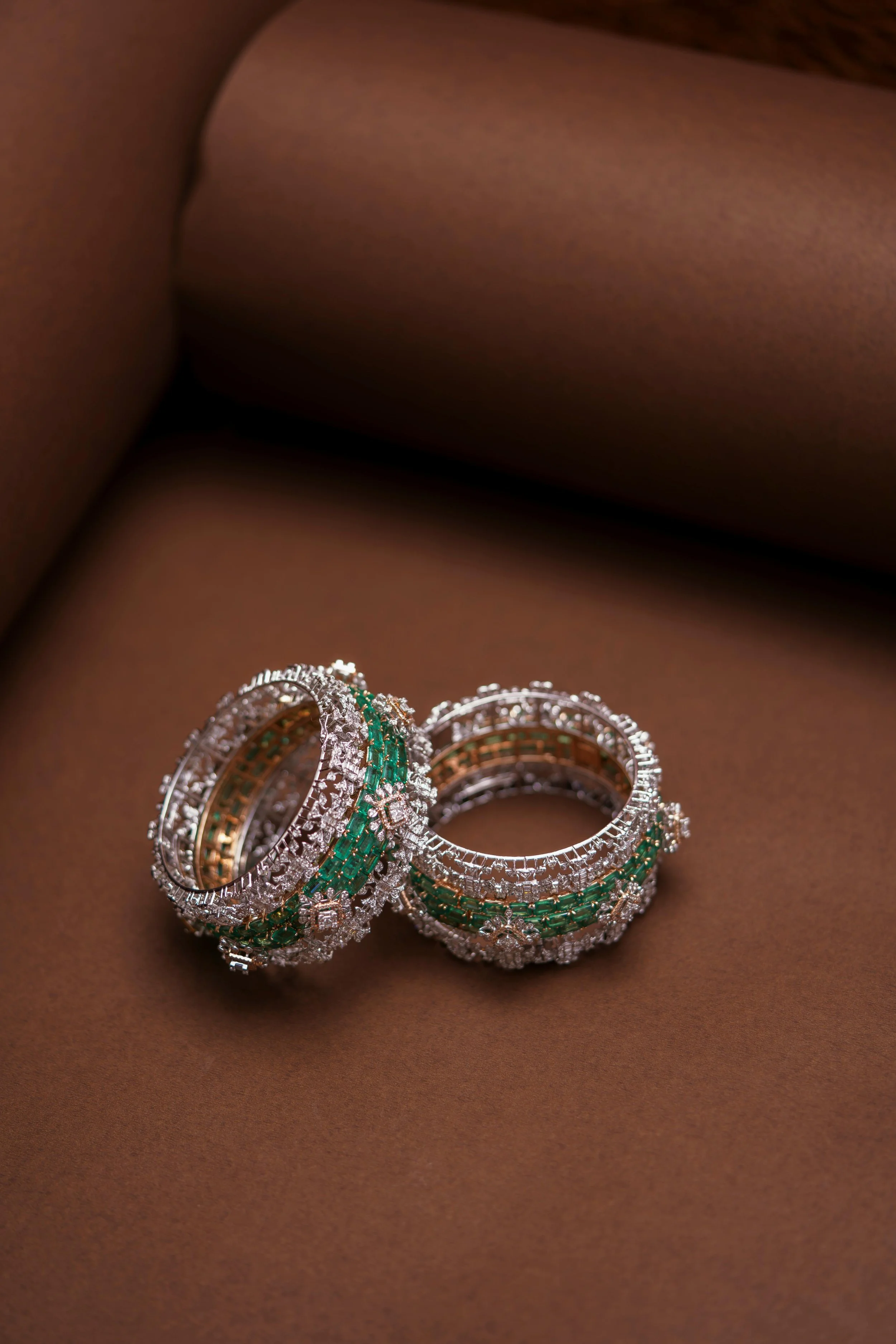 Two ornate rings decorated with diamonds and emeralds resting on a brown surface.