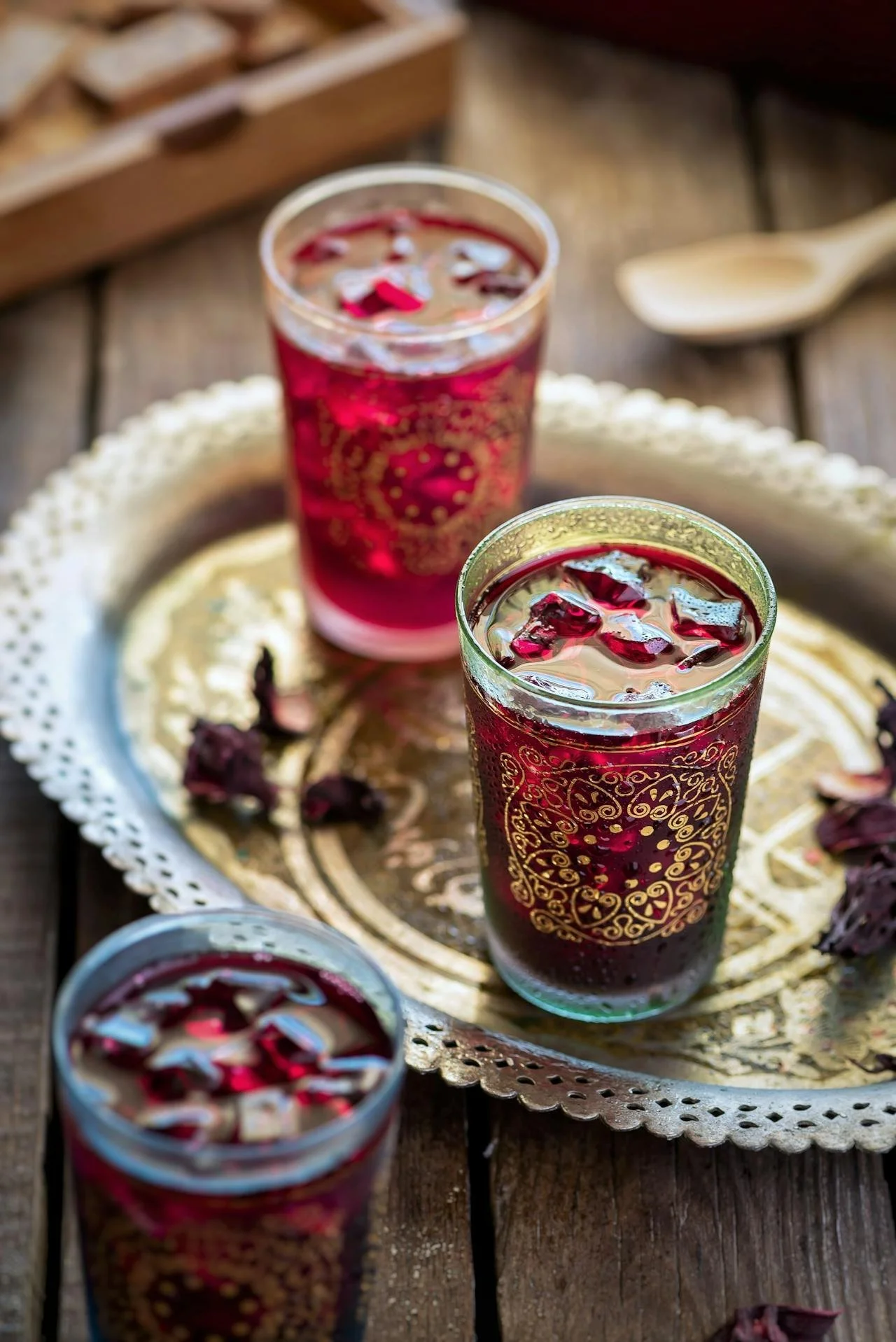 Three glasses of a dark red beverage with ice and rose petals on a decorative silver tray on a wooden table.
