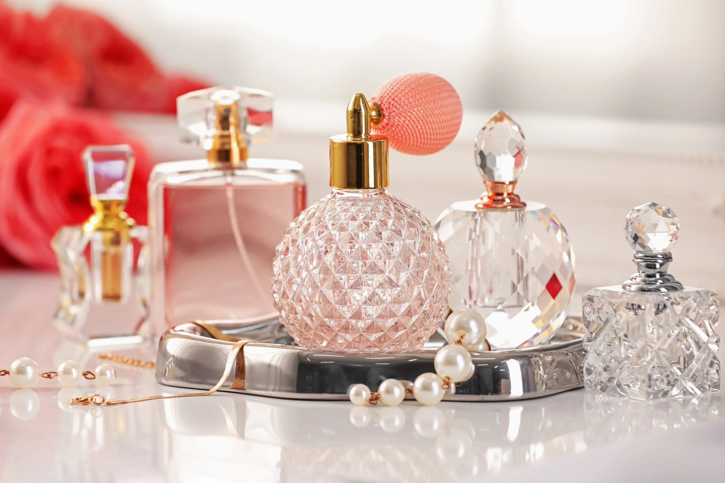 Luxury perfume bottles with crystal designs and decorative pearls, set on a silver tray with a soft pink background including pink roses.