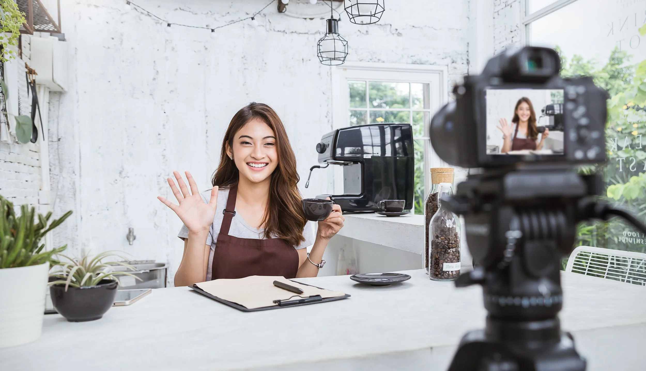 Young woman in apron waving and smiling at camera in a bright coffee shop, with coffee machine, plants, and clipboard on table, camera recording her.