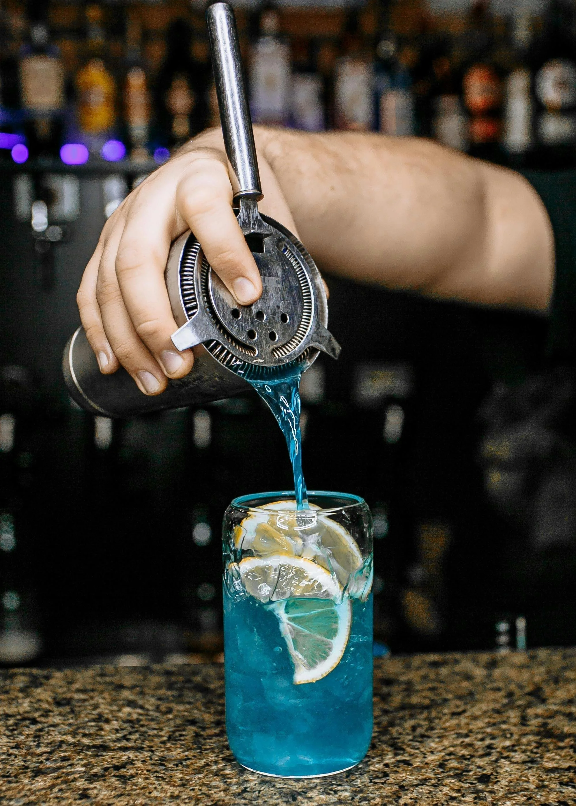 A bartender pours blue liquid from a cocktail dispenser into a tall glass with lemon slices and ice.