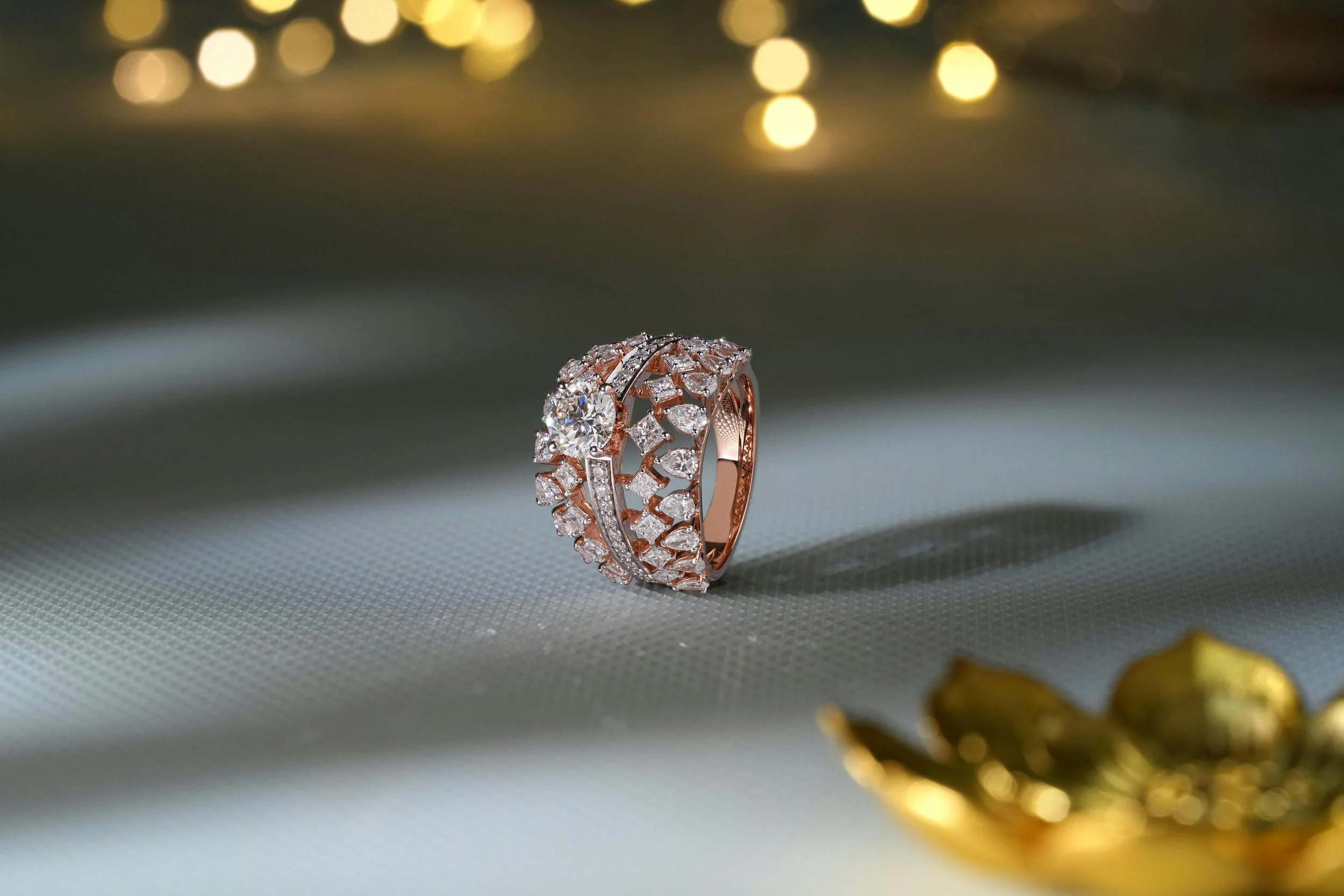 A gold and silver diamond ring with intricate design, placed on a textured gray surface with blurred golden background lights.