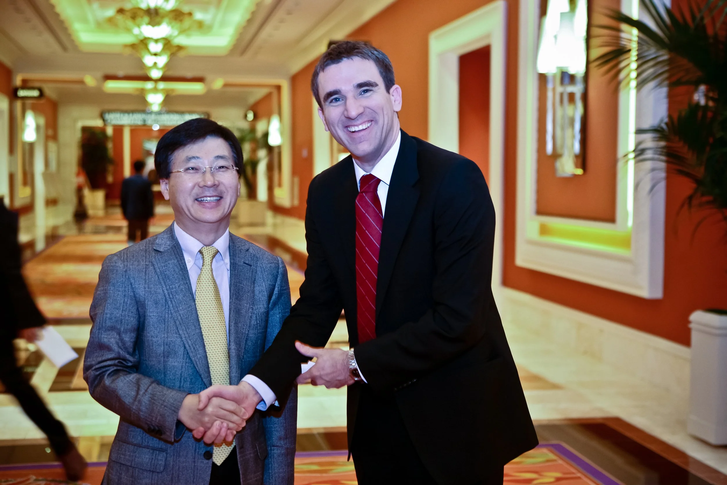 Two men in suits shaking hands and smiling in a well-lit hotel corridor with orange walls and decorative lighting.
