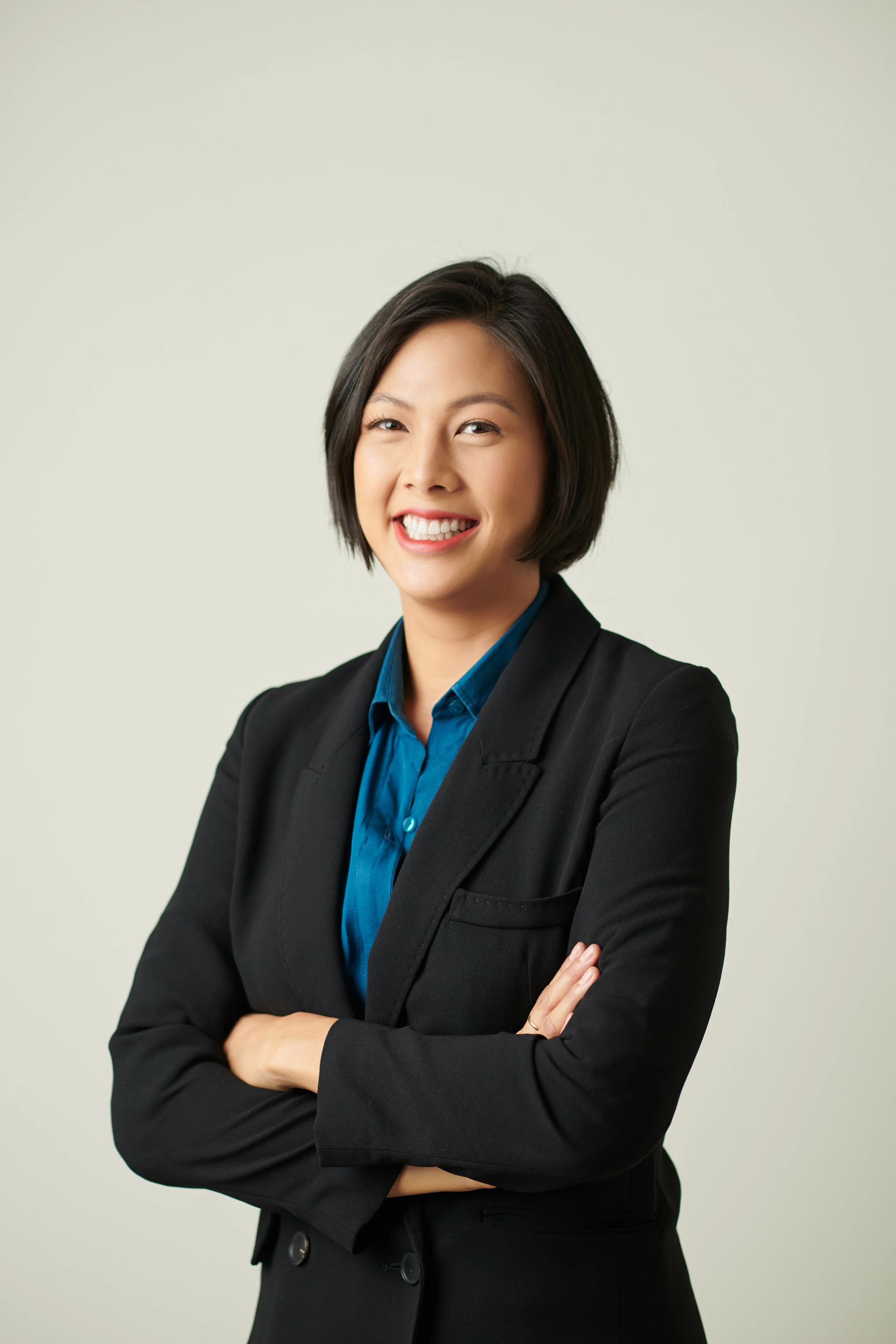 Professional woman with short black hair smiling, wearing a black blazer and blue shirt, crossing her arms against a plain light background.