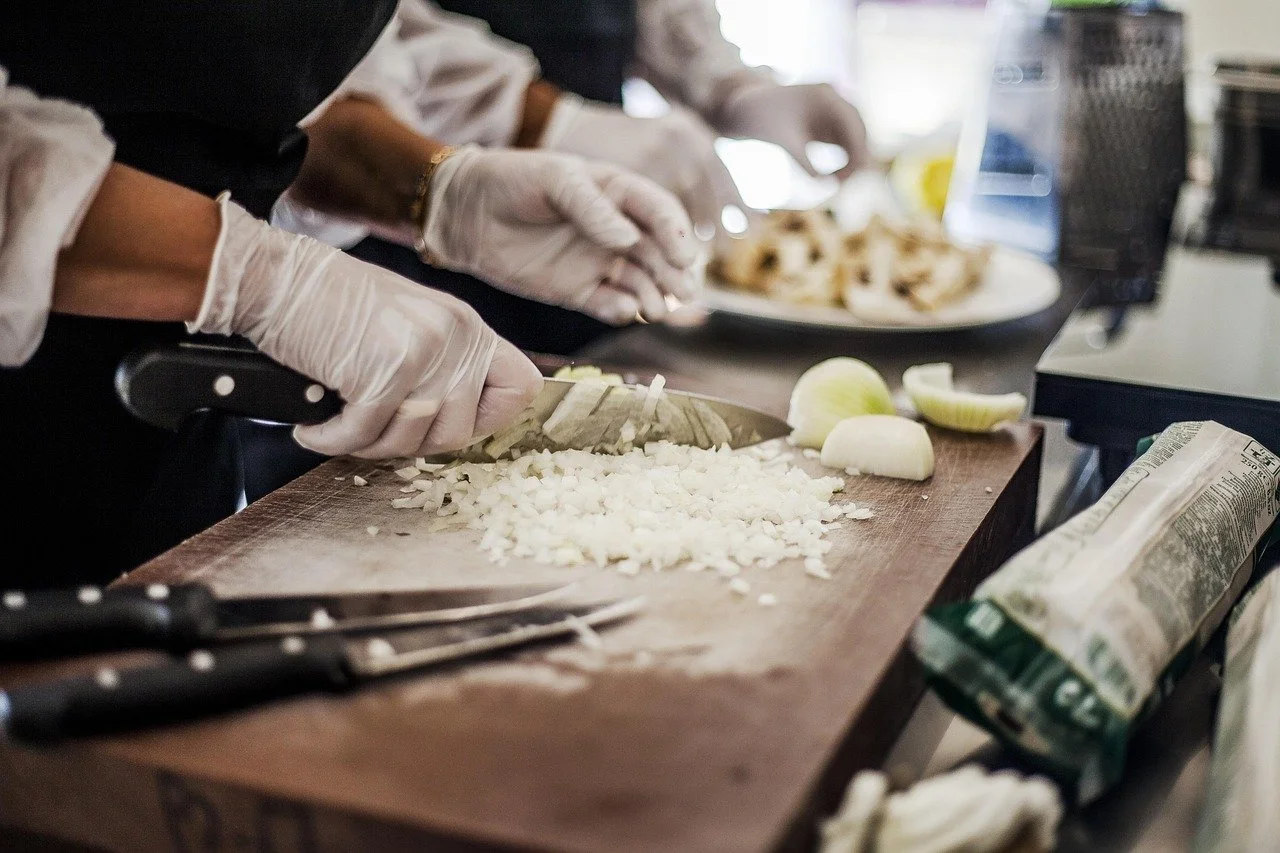 Close-up of people chopping onions on a wooden cutting board in a kitchen, with food preparation tools and ingredients nearby.