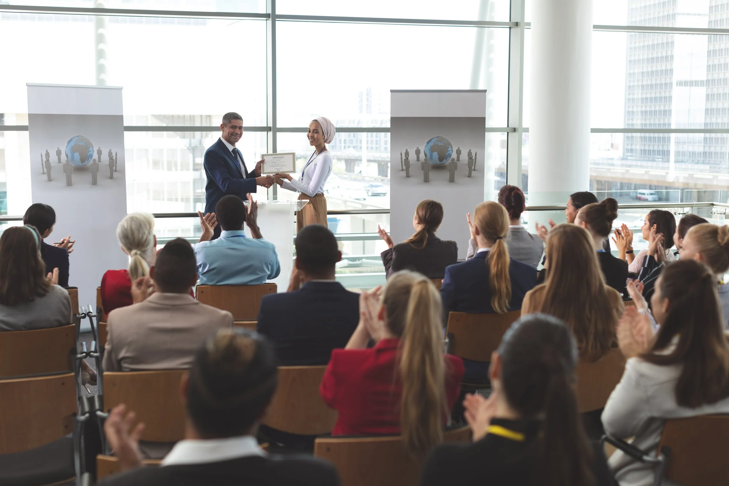 A woman receiving a certificate from a man during an award ceremony in a modern conference room with large windows, with an audience applauding.