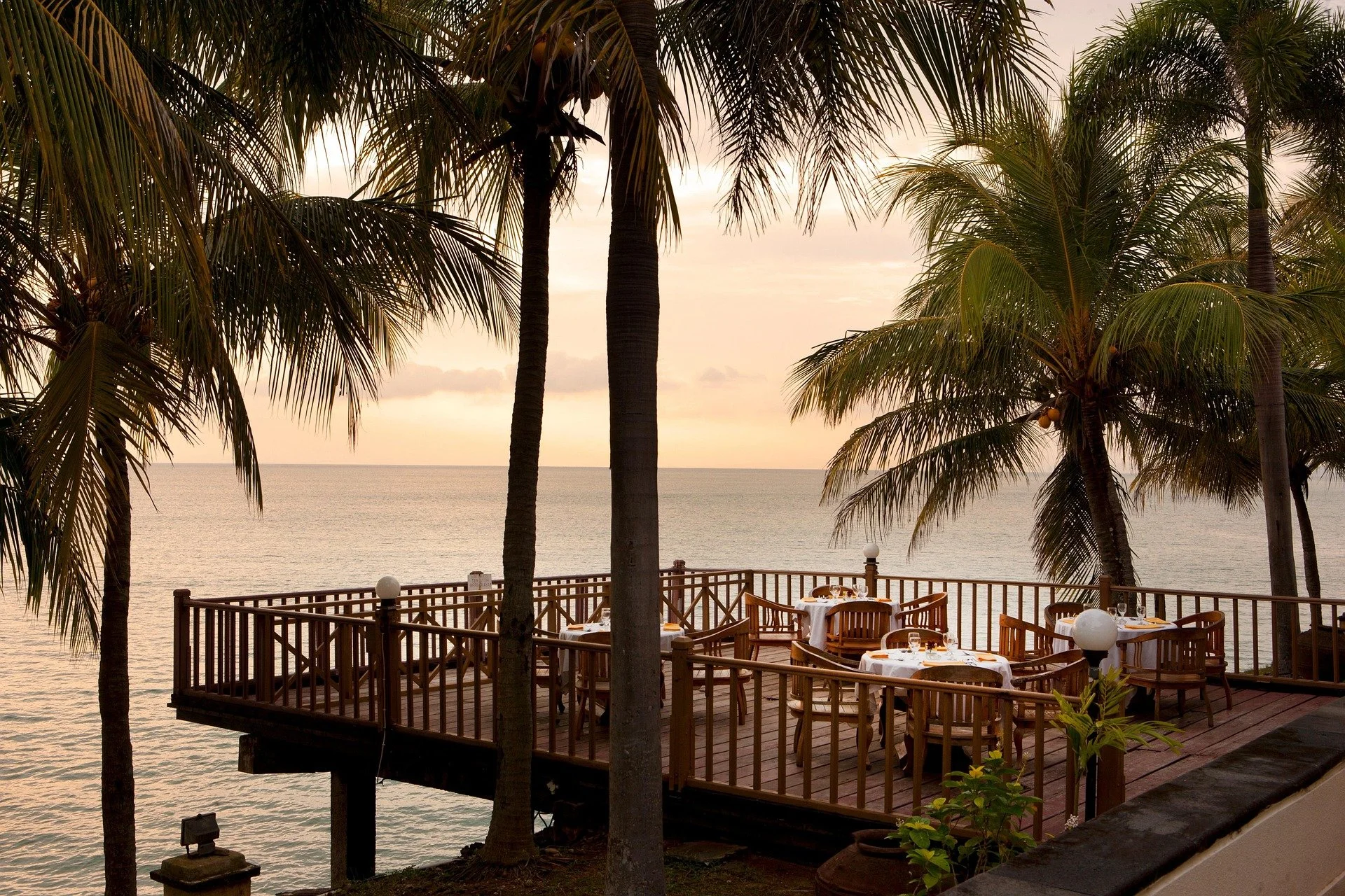 Outdoor dining area on a wooden terrace overlooking the ocean at sunset, with palm trees surrounding the space