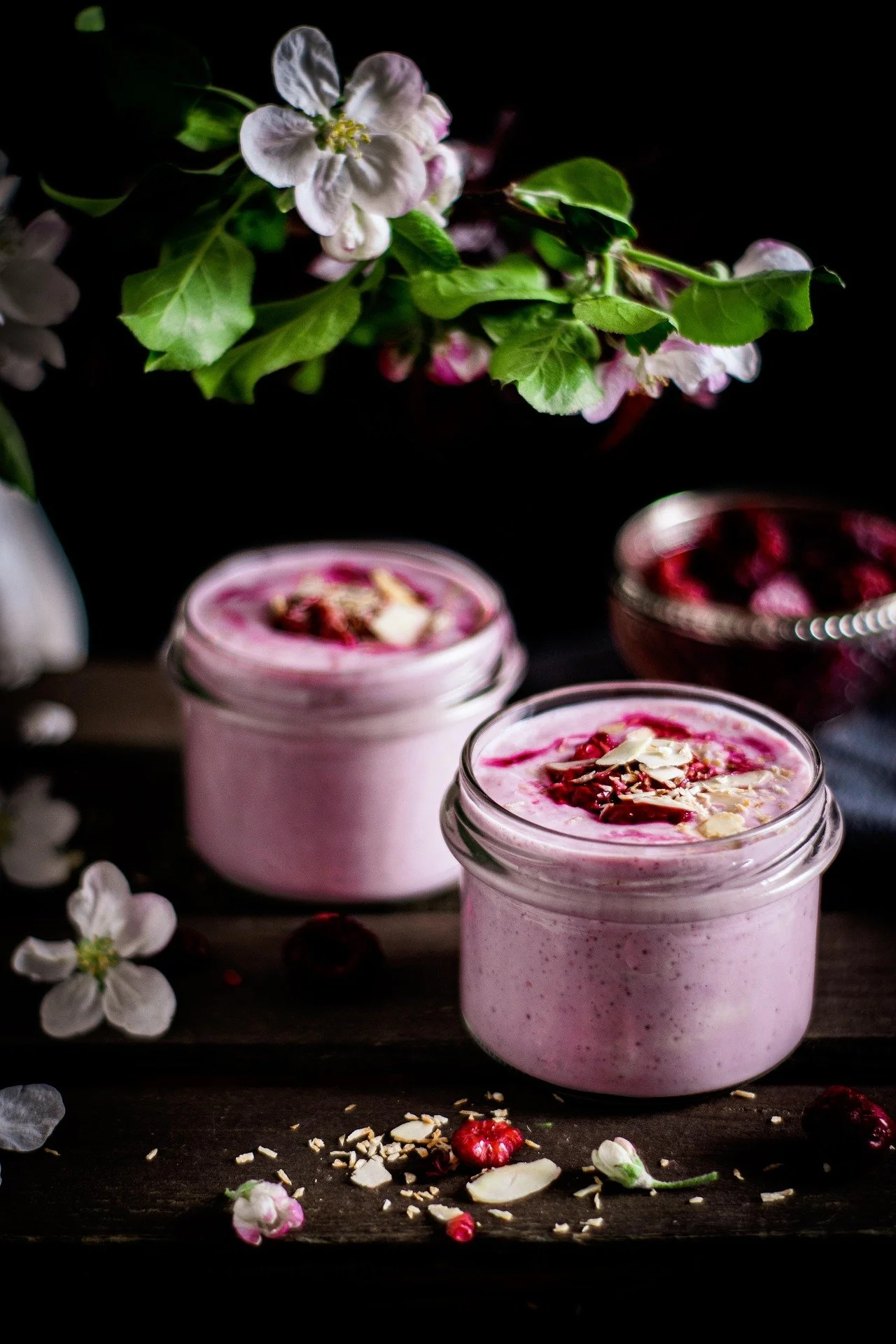 Two jars of pink yogurt topped with berries and almond slices on a dark wooden surface, with white flowers and green leaves in the background.