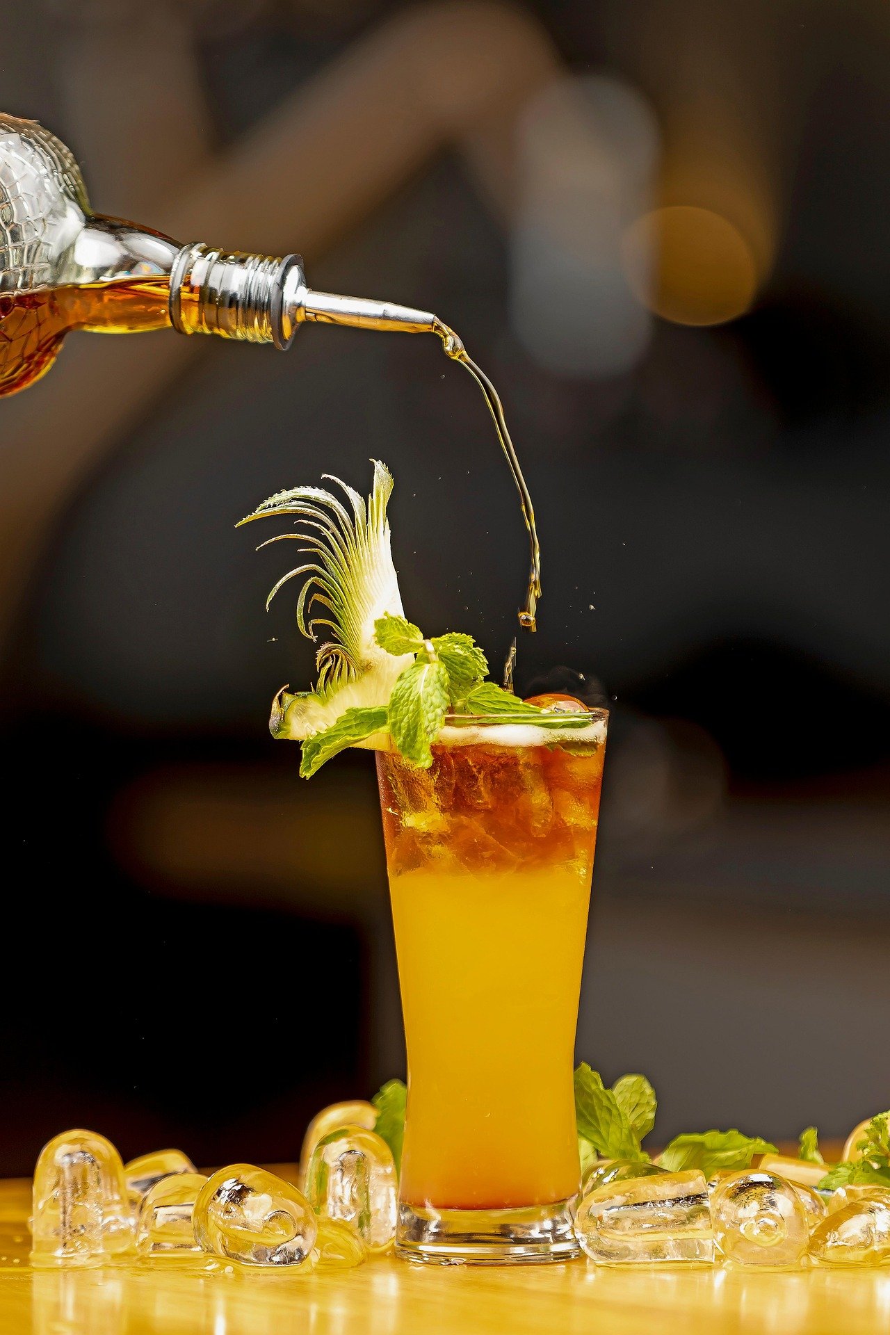 A tropical cocktail garnished with a lime wedge, mint leaves, and a feathery vegetable, with a splash of alcohol being poured into it, surrounded by ice cubes and mint leaves on a wooden surface.