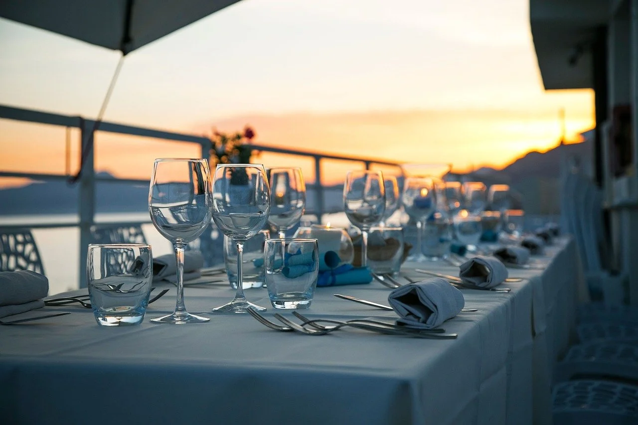 Elegant outdoor dining table set for a meal during sunset with wine glasses, water glasses, silverware, napkins, and a floral centerpiece, overlooking water and mountains.