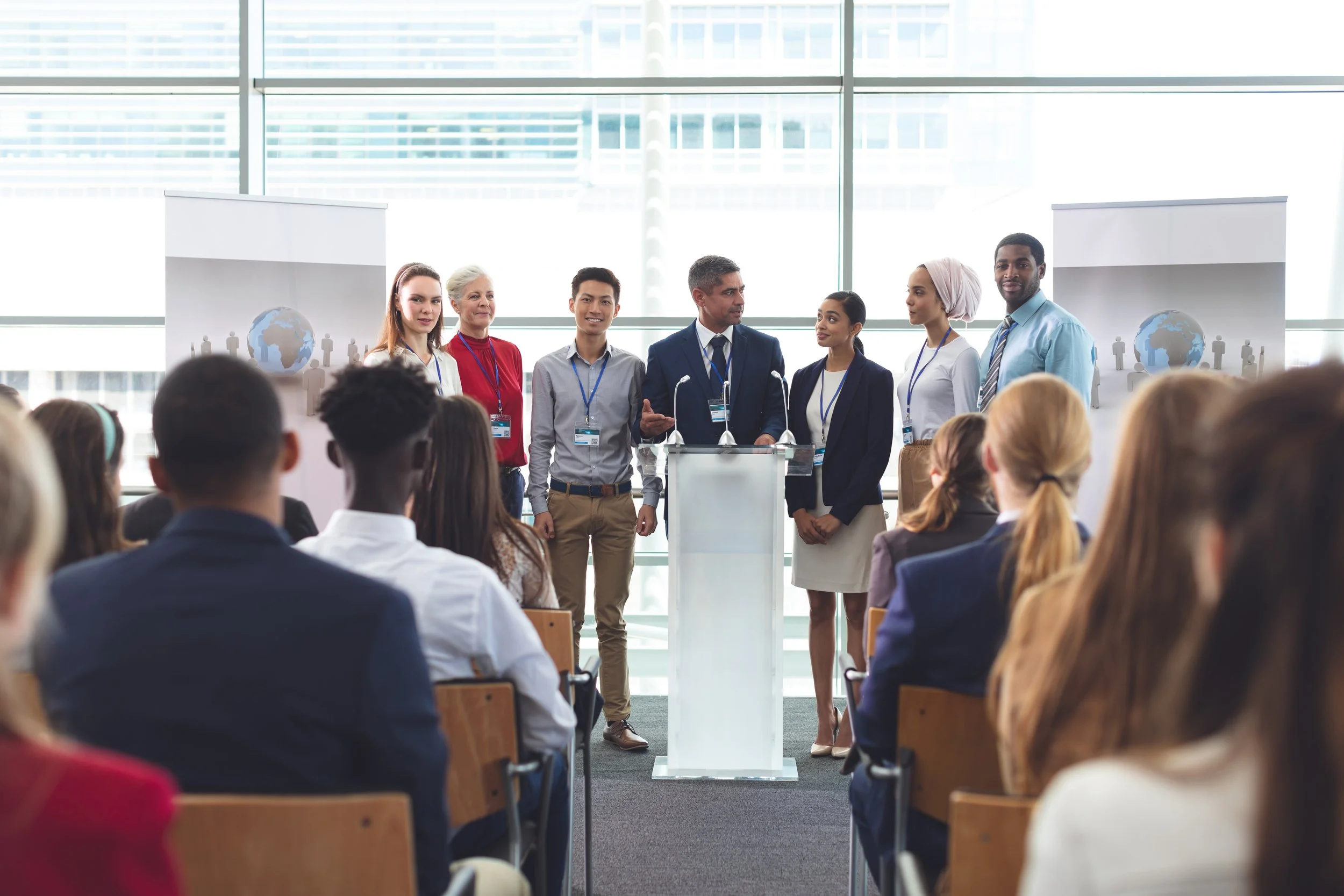 A diverse group of speakers standing at a podium during a conference or presentation in a modern, glass-walled room, with an audience seated facing them.
