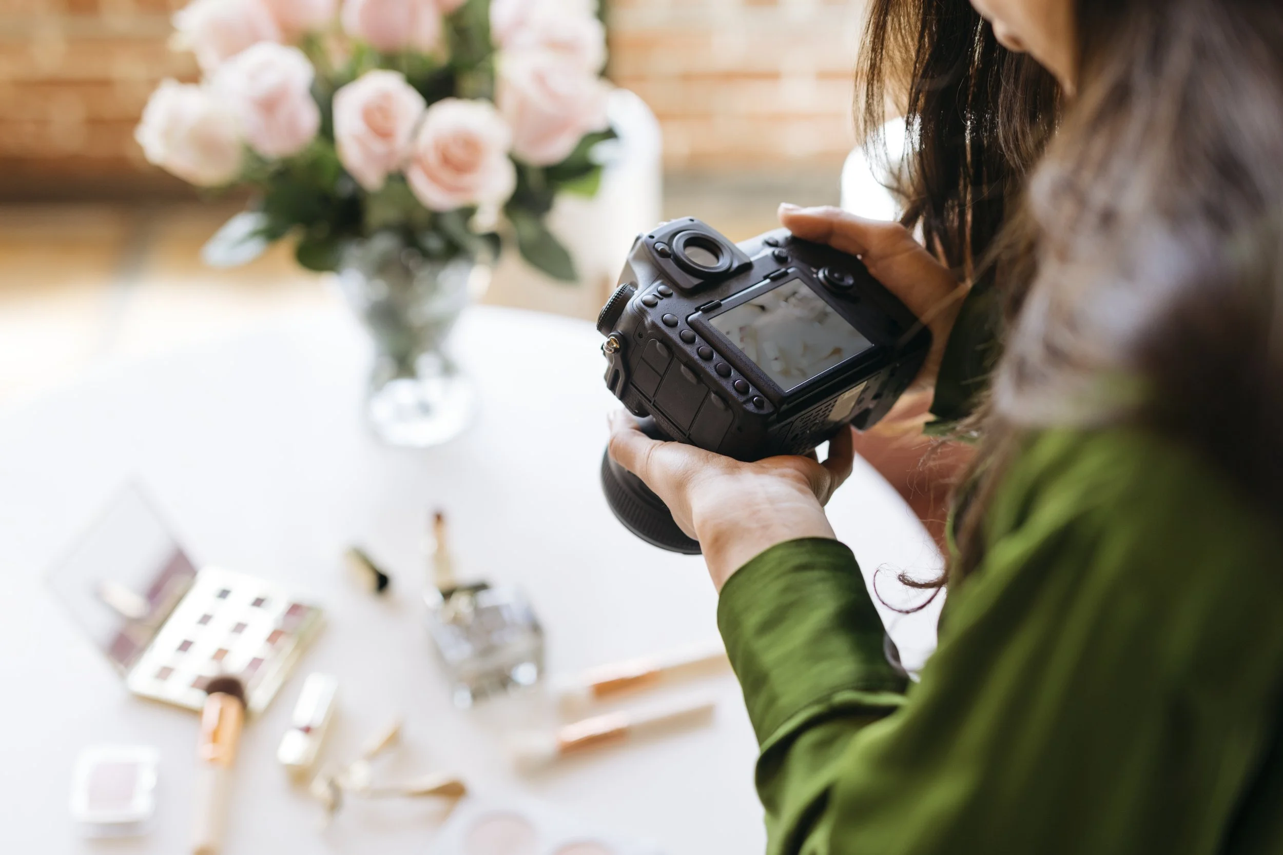 A woman in a green shirt is holding a camera and taking a photo of a table with makeup products and a vase of pink roses.
