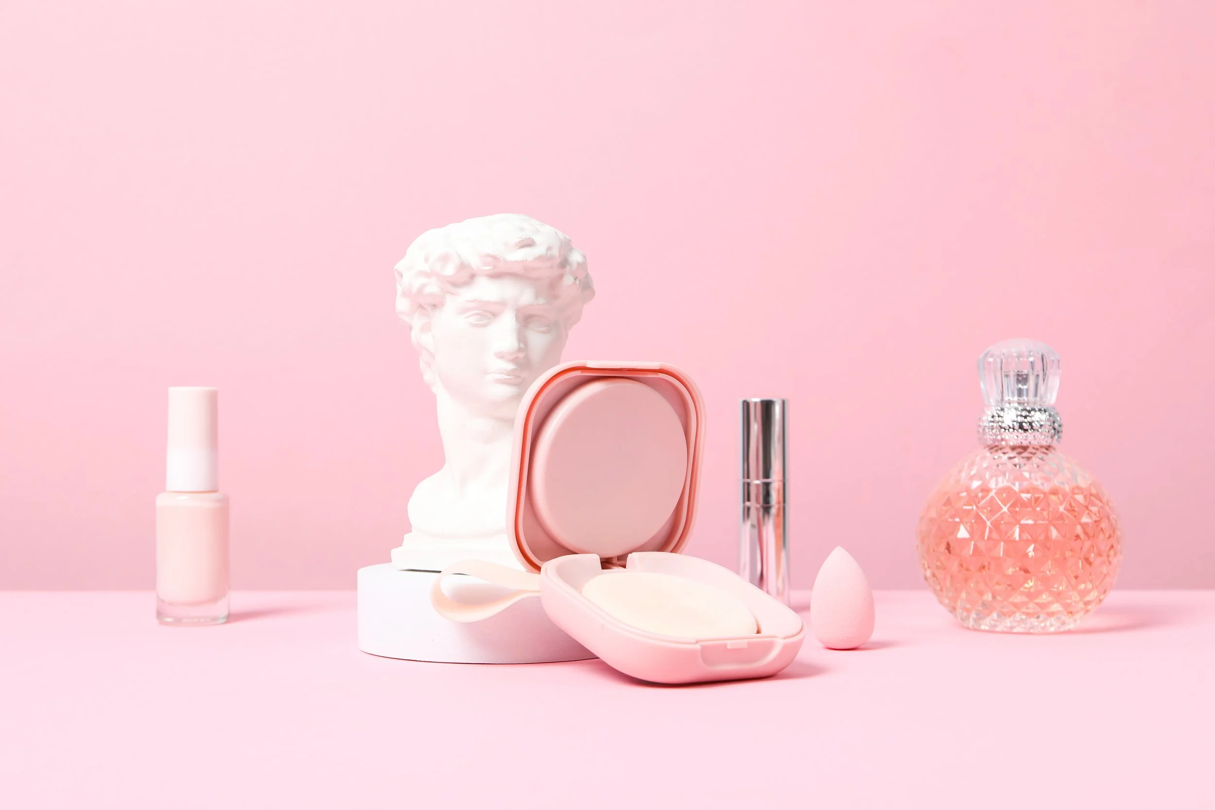 Pink background with skincare products, a nail polish, a lipstick, a perfume bottle, a compact mirror, a powder puff, a makeup sponge, and a white bust sculpture of a classical male figure.