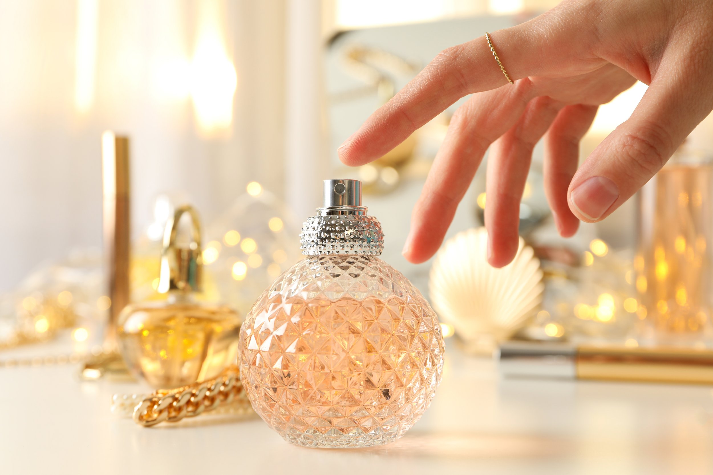 A person's hand reaches towards a decorative perfume bottle with a textured, diamond-patterned surface, surrounded by other perfume bottles and jewelry on a vanity or dressing table.