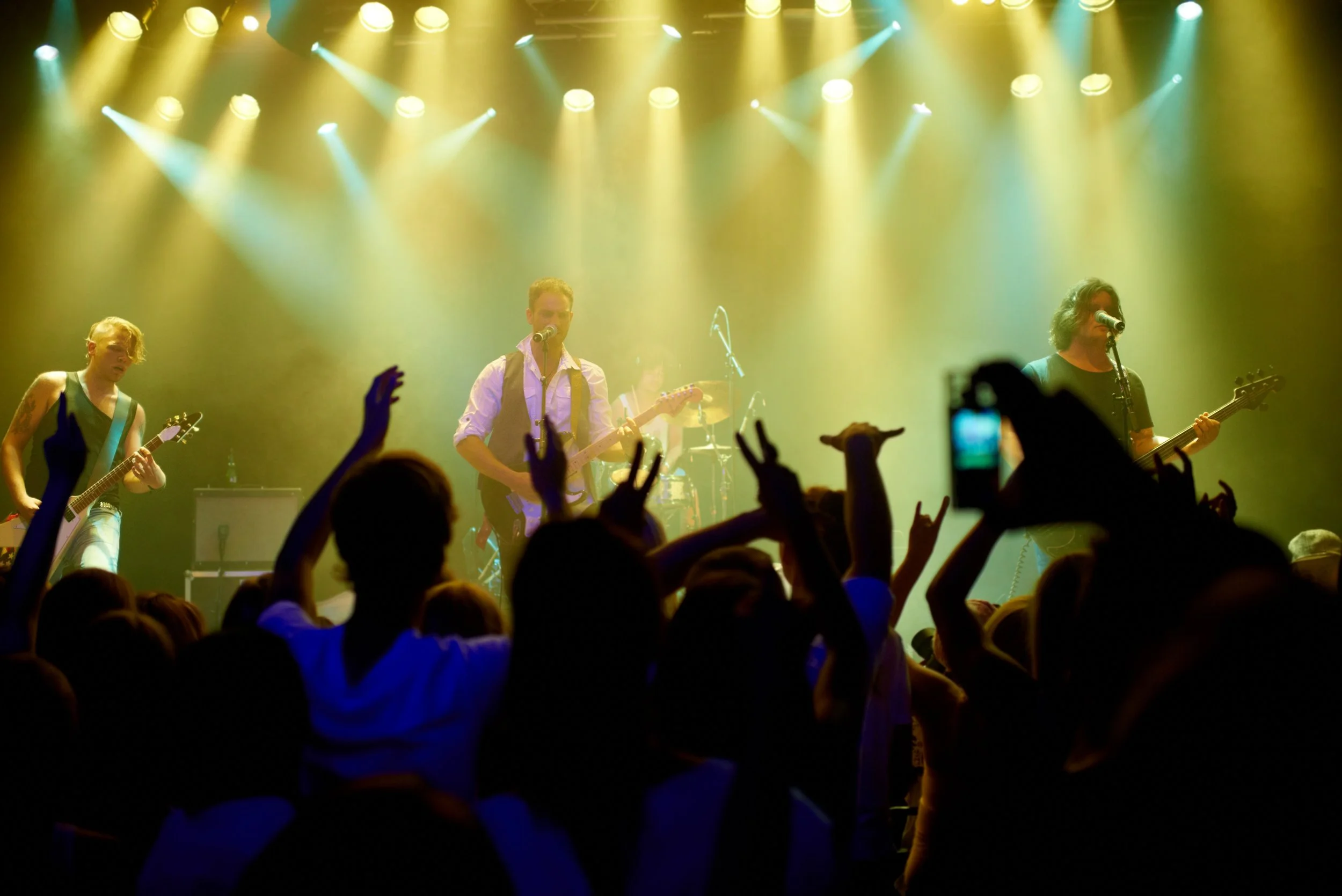 A band performing live on stage with yellow concert lights, playing electric guitars and drums, while an audience dances and raises their hands.