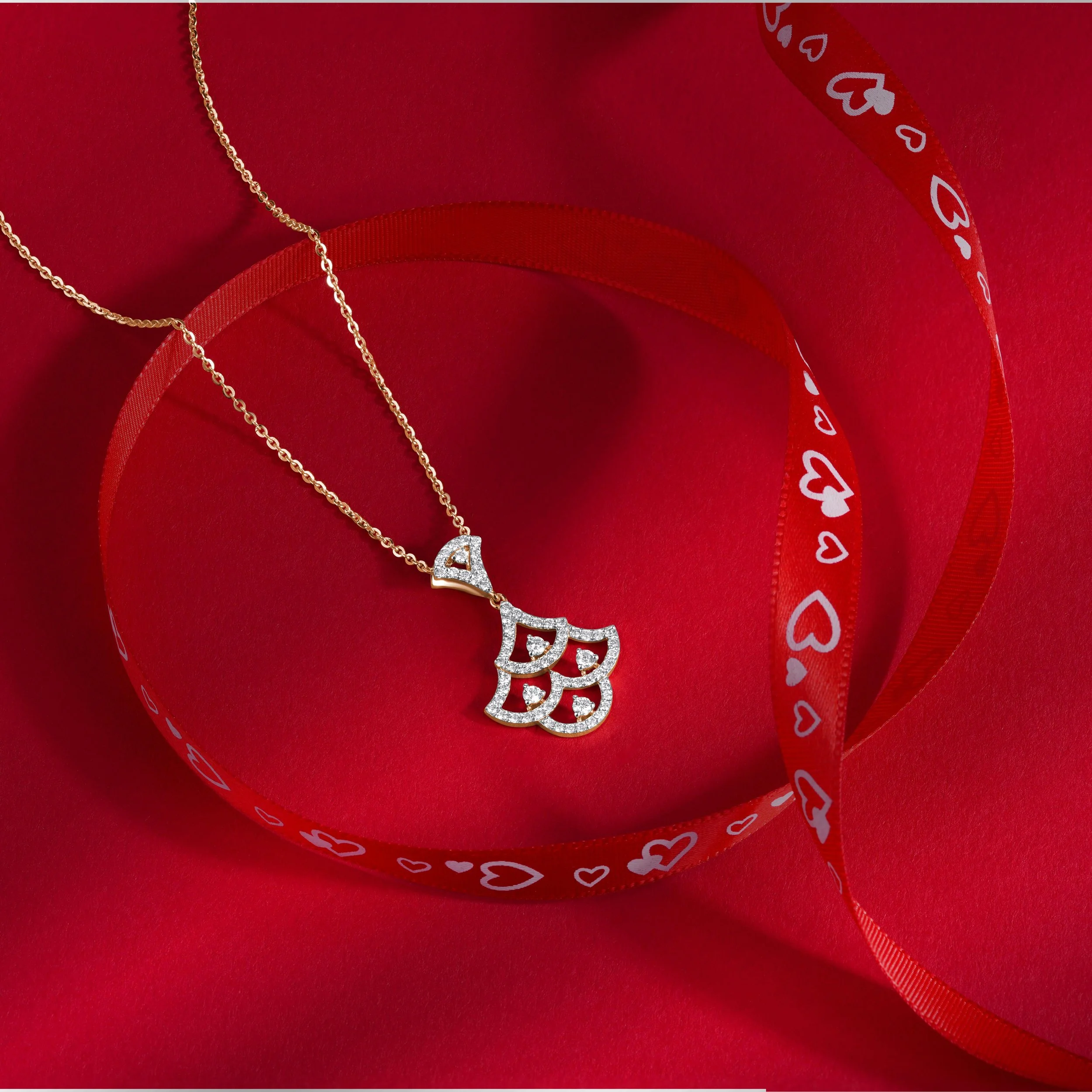 Gold necklace with a pendant featuring three heart designs and the letter 'B', decorated with small diamonds, placed on a red background with a red heart-shaped ribbon with white hearts and the letter 'B' printed on it.