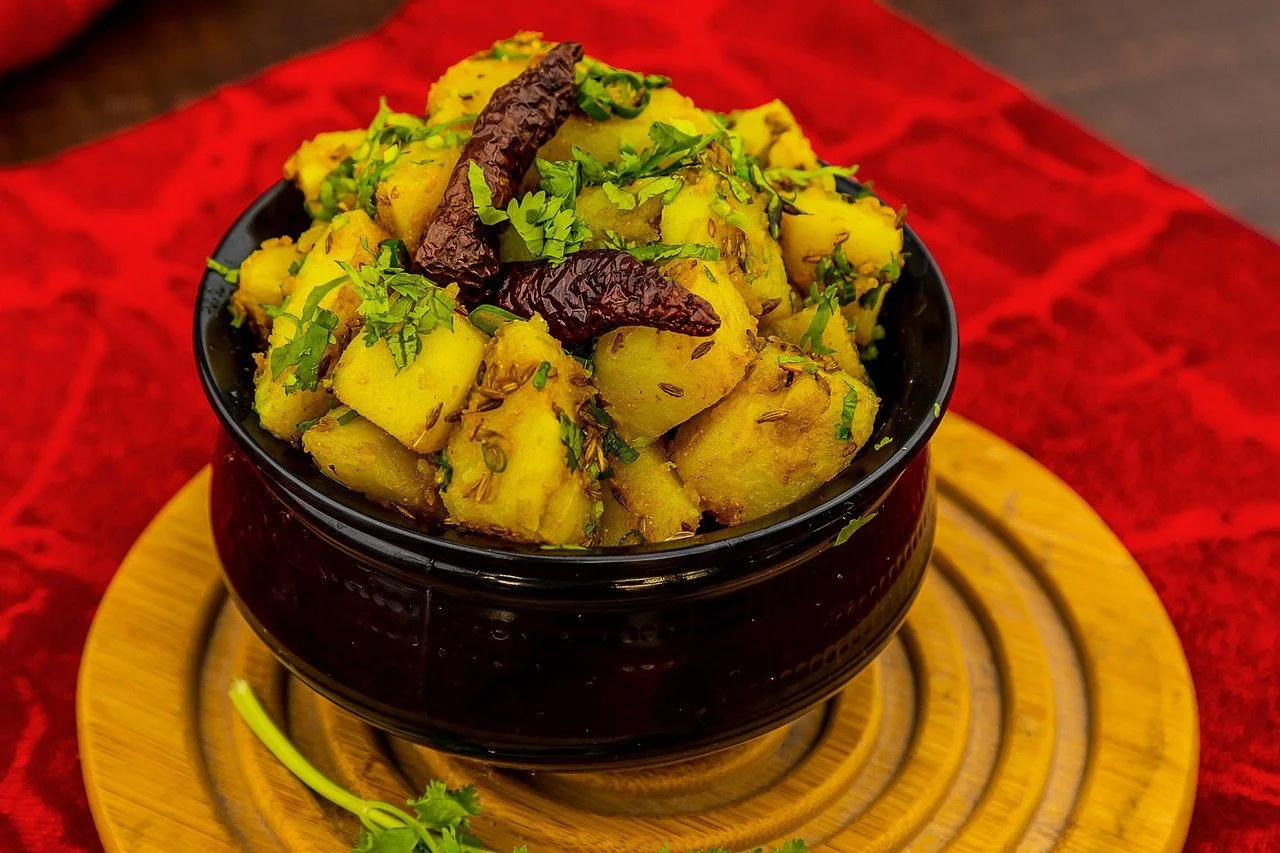 Bowl of seasoned potatoes garnished with cilantro and dried red chili peppers, placed on a wooden serving board with a red cloth underneath.