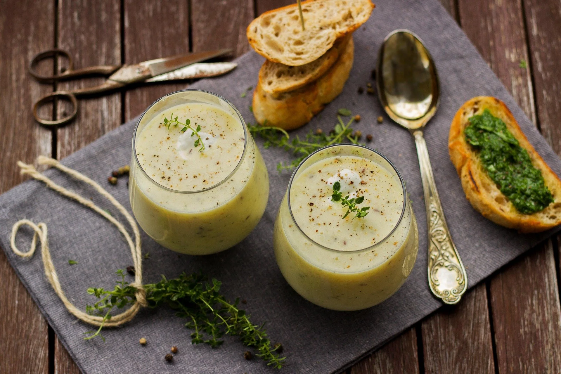 Two glasses of creamy soup garnished with herbs on a gray cloth, with slices of bread and a roll filled with green spread, set on a wooden table.