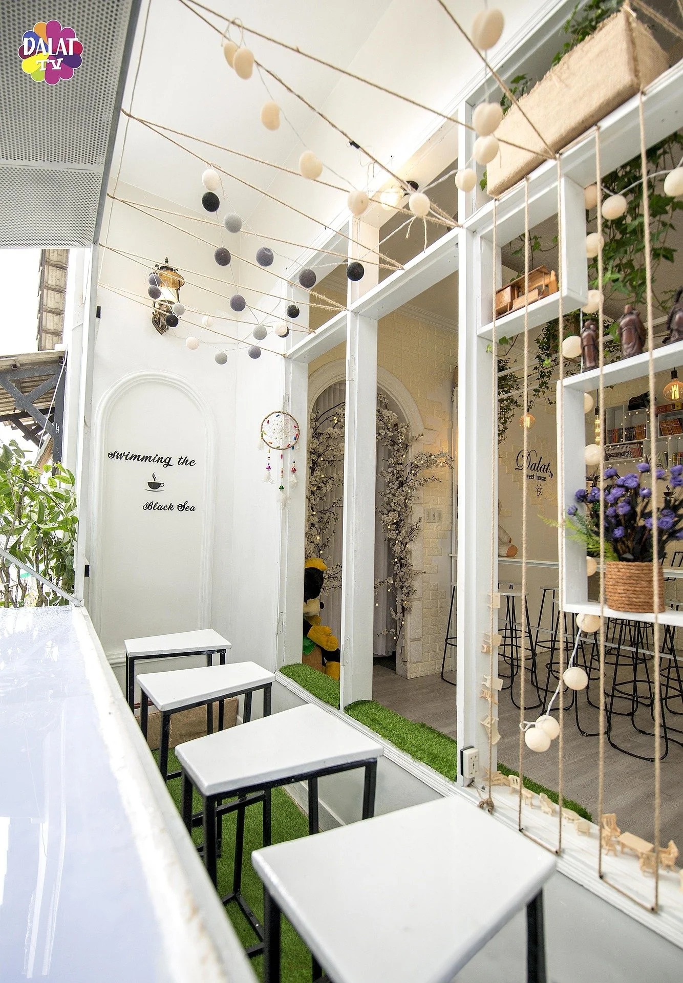 Indoor cafe with string lights, decorative felt ball garlands, potted plants, bar stools, and white tables with black legs.