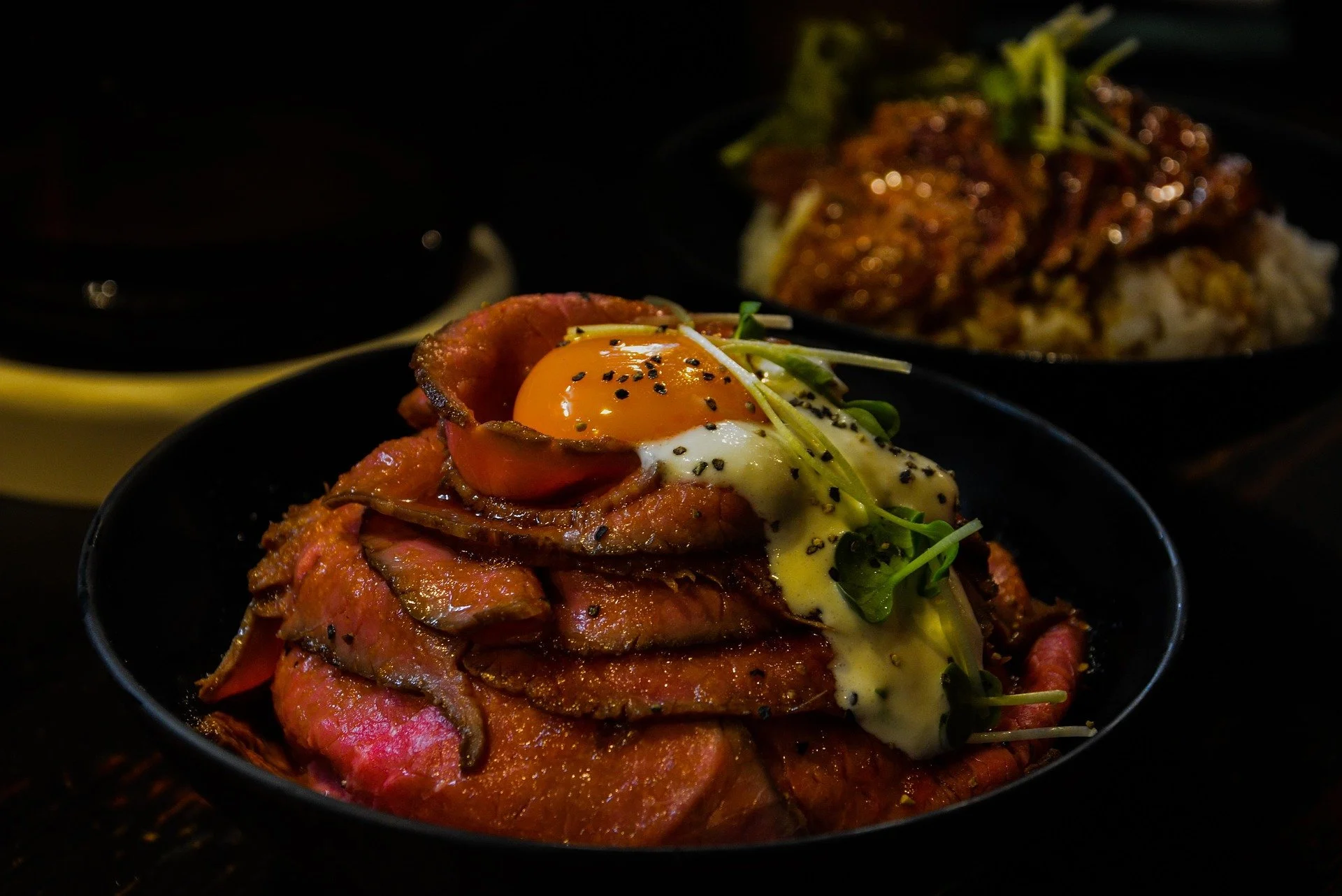Bowl of sliced rare roast beef topped with a fried egg, microgreens, and black pepper. In the background, a bowl of rice and another dish of meat and vegetables.