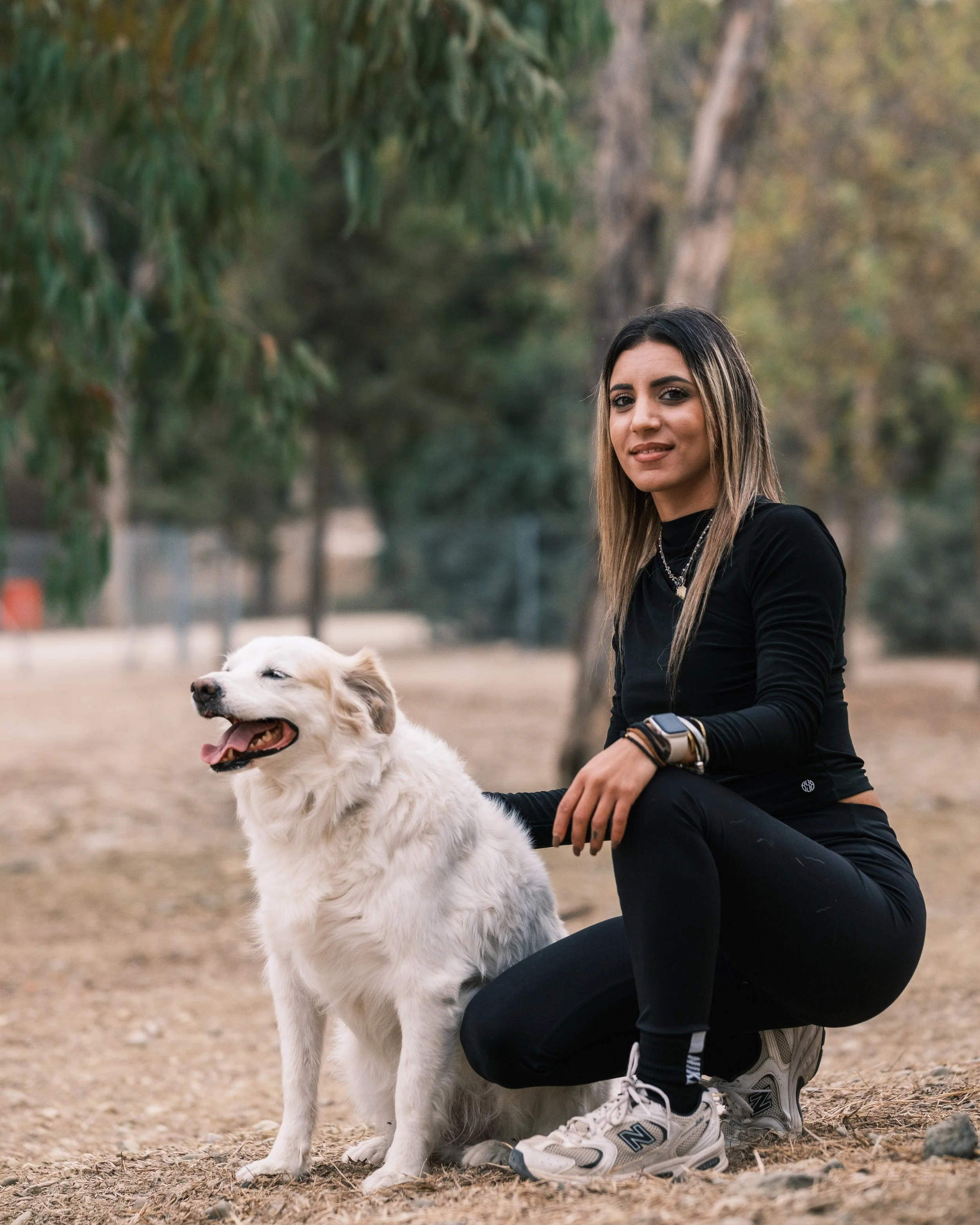 A woman kneels next to a happy, white and beige dog in a park with trees in the background.