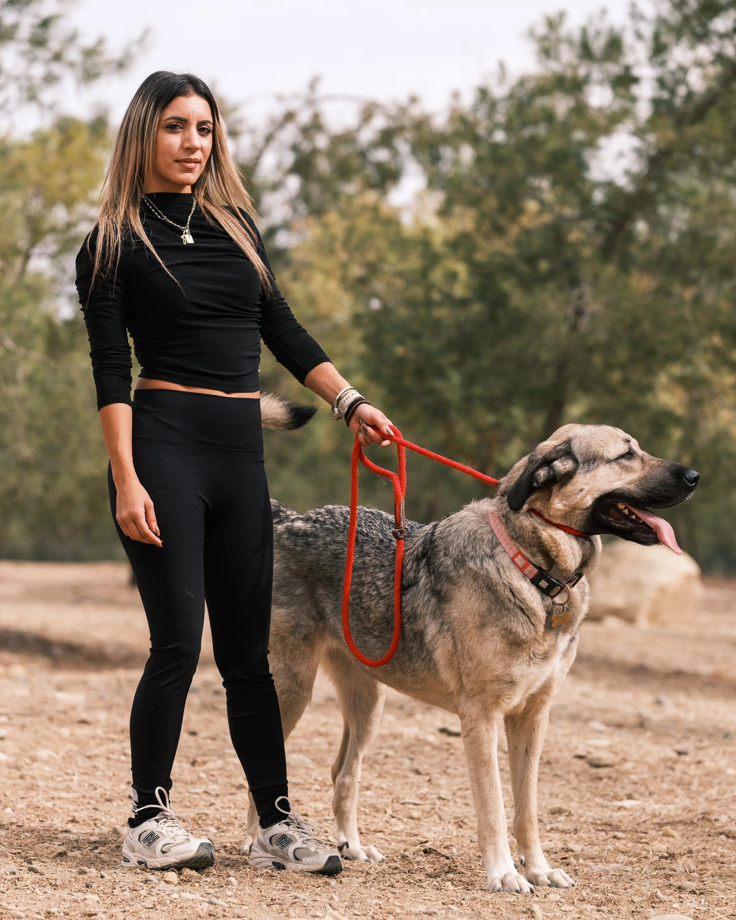 A woman standing outdoors with a large dog on a leash, in a park or wooded area.