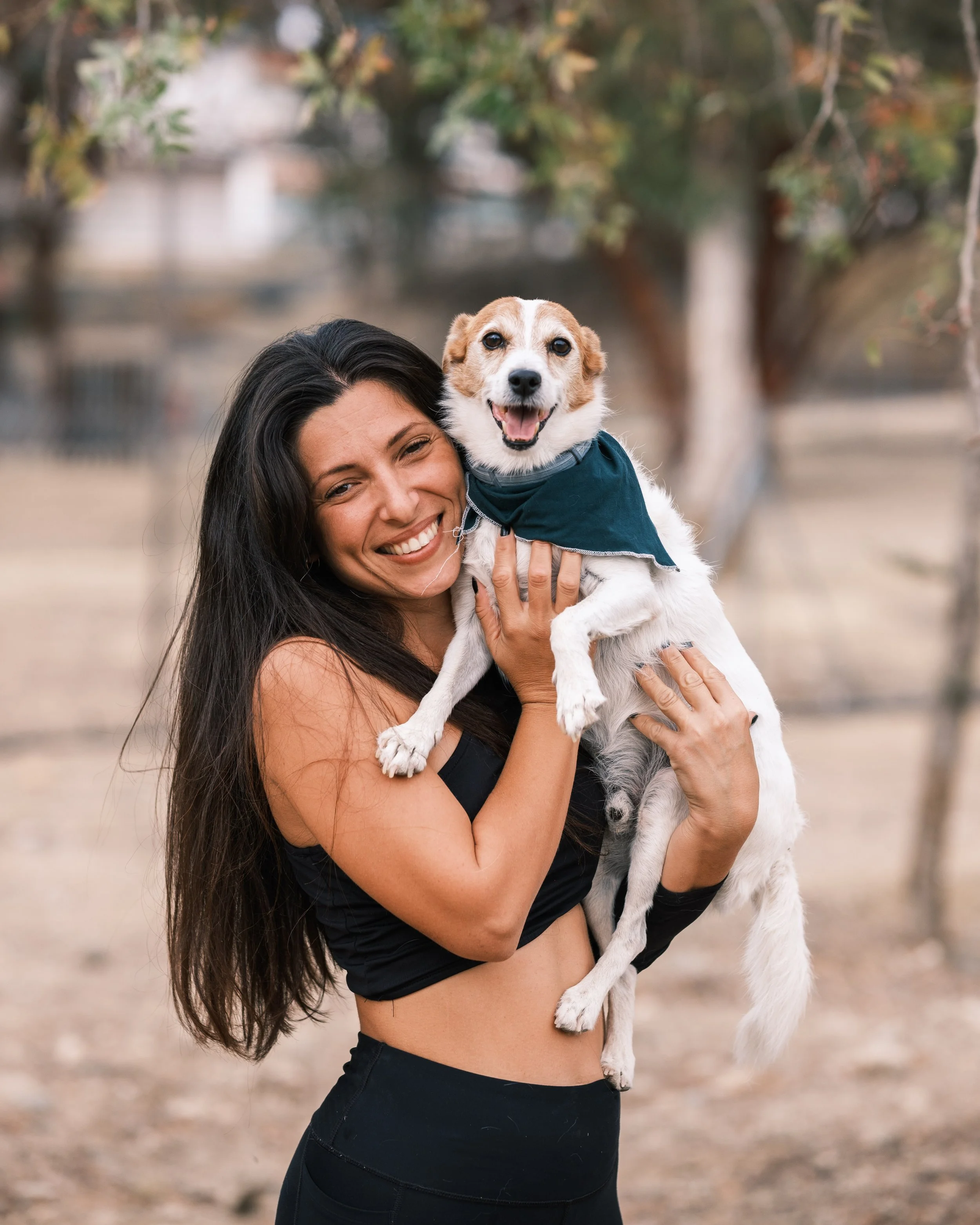A woman with long dark hair smiling and holding a small dog with light brown and white fur, wearing a dark bandana, outdoors with trees in the background.