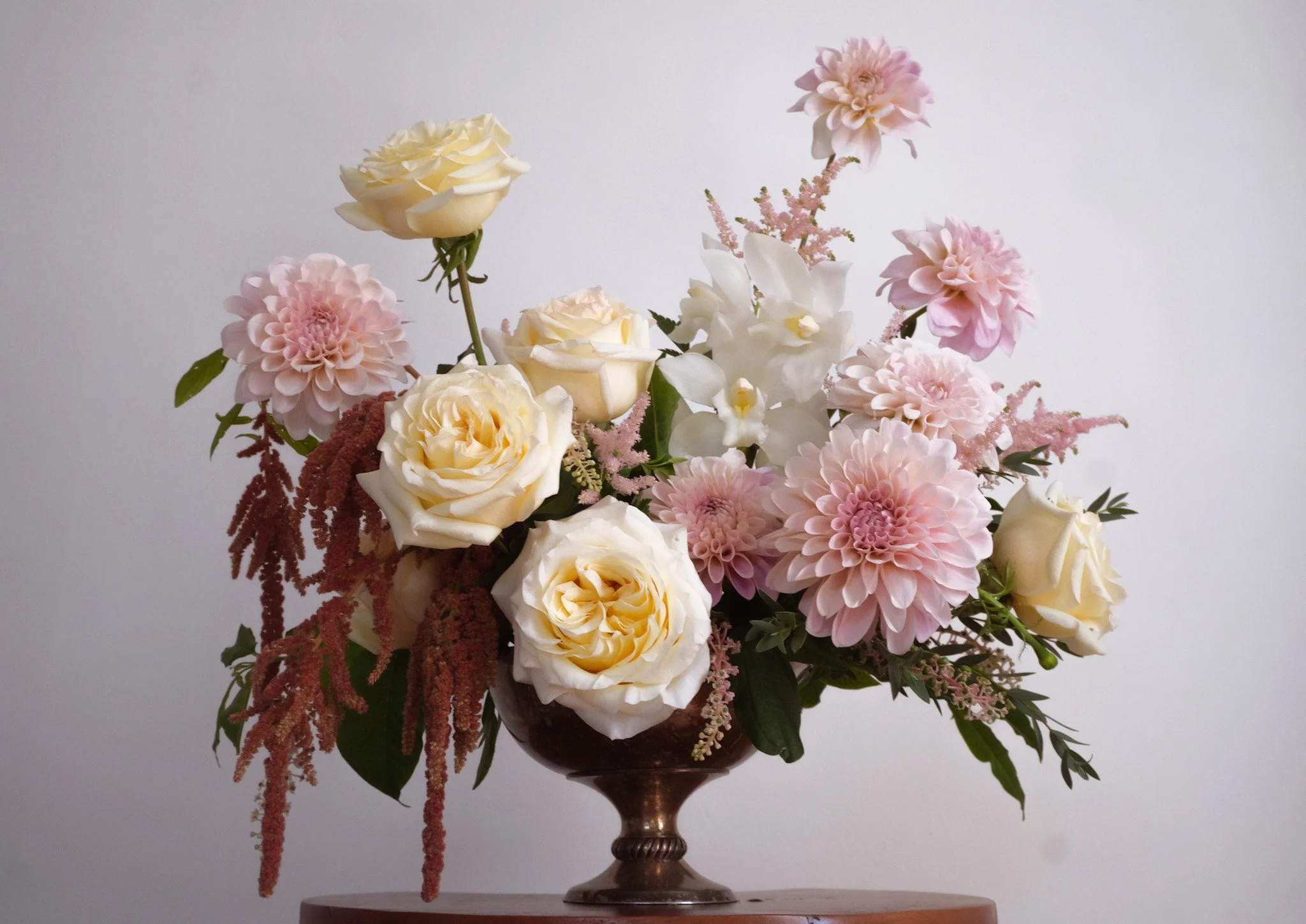 A flower arrangement in a dark bronze vase containing white roses, pink dahlias, white orchids, pink astilbe, and green foliage against a plain light background.